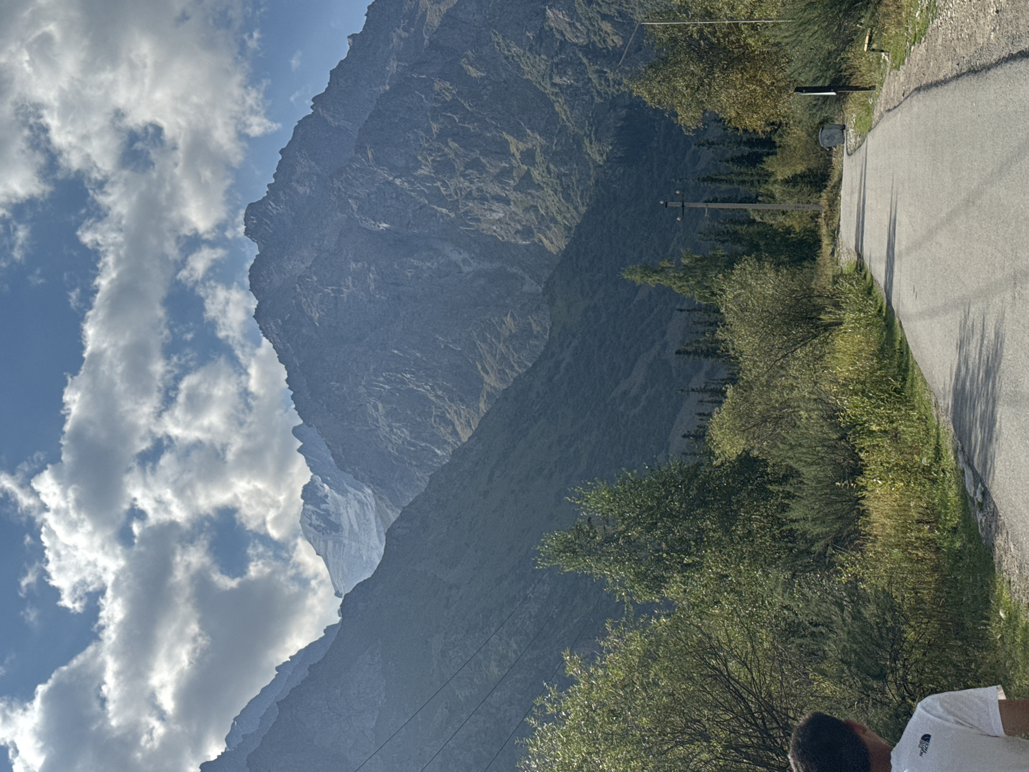 A winding asphalt road curves through a grassy landscape toward a dense forest and towering, rugged mountains under a cloudy blue sky. A person wearing a white t-shirt is partially visible on the left side of the frame, facing away from the camera and towards the road. The road is bordered by green brush and trees on either side, with a power line pole and a trash bin visible in the distance. The mountains in the background are covered in greenery at their base and appear rocky and snow-capped at their peaks. The scene is bathed in natural sunlight, casting shadows across the road and vegetation. The location is identified as being in the vicinity of Ala-Archa National Park, Kyrgyzstan.