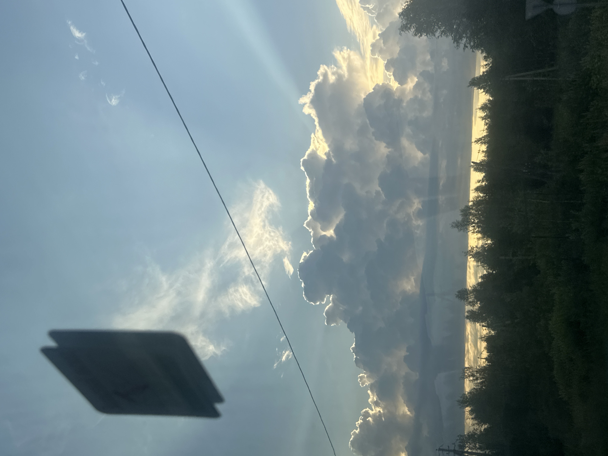 A partially obscured, dark rectangular object, possibly a sign or sticker, is affixed to a glass surface, casting a shadow. Beyond this surface, a wide expanse of the sky dominates the frame, featuring a blend of light blue and white clouds. Dramatic sunbeams pierce through the clouds, illuminating them from behind and creating a radiant halo effect. A thin, dark horizontal line, likely a wire, stretches across the upper portion of the sky. The lower third of the image reveals a dense treeline of varying heights, with a strip of hazy, yellow light visible above the trees. The ground in the foreground is covered in dark vegetation. The scene appears to be captured outdoors, likely from a moving vehicle given the perspective and the presence of the object on the glass. The location is specified as Yarkovo, Russia.