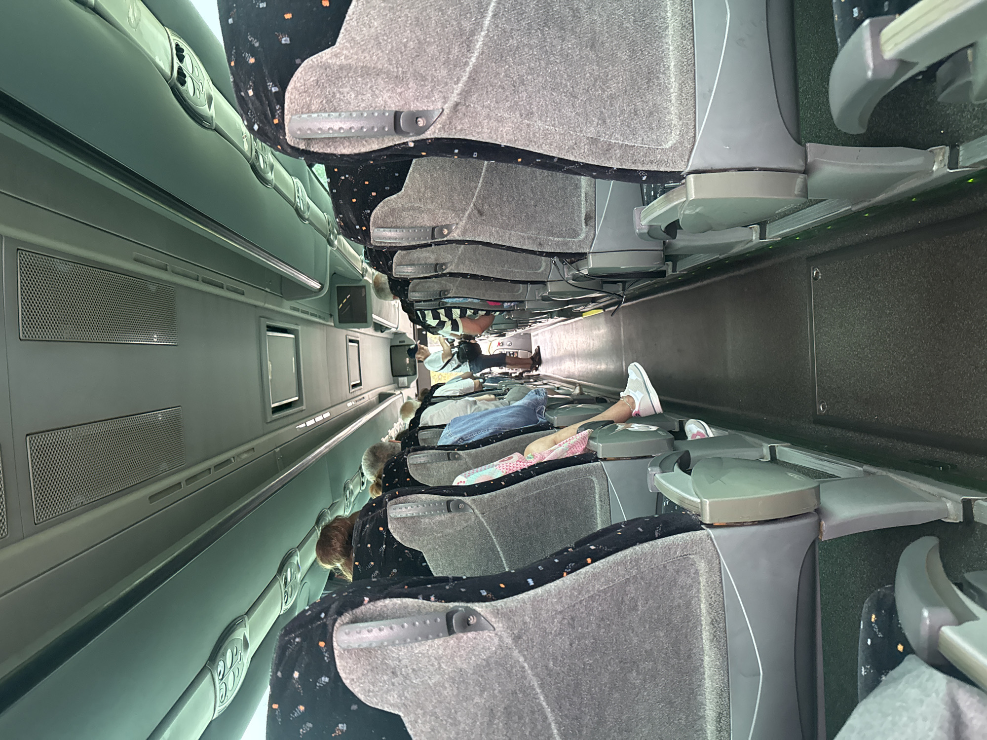 The image shows the interior of a tour bus, likely in Fuengirola, Spain, with passengers seated. The viewpoint is from one of the seats, looking down the aisle of the bus. On the right, a row of grey fabric seats with a black pattern of small orange and white dots is visible. Each seat has grey plastic armrests and supports. The aisle floor is dark grey. On the left, the back of a grey fabric seat with the same black pattern is partially visible. In the foreground on the left, the back of another seat and a glimpse of a person's legs are seen. Further down the aisle, several passengers are visible, some with their backs to the camera and others seen from the side. One passenger in the distance is standing and appears to be talking to someone. The ceiling of the bus is light grey and features air vents and lighting fixtures. A small screen is mounted on the ceiling towards the front of the bus. The overall impression is of a typical bus interior, suggesting travel or a guided tour.