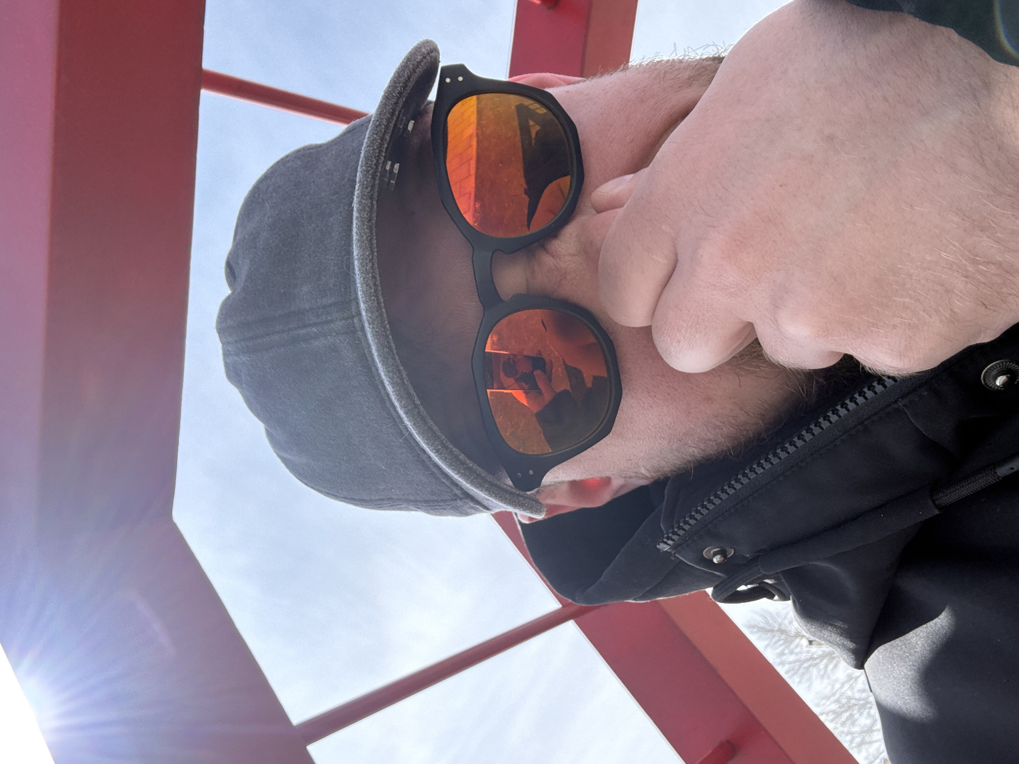 A low-angle close-up shot depicts a person wearing a dark grey baseball cap and black-framed sunglasses with orange reflective lenses. The person is also wearing a black jacket. The individual's right hand is partially obscuring their face. Reflected in the sunglasses are red architectural elements. The image also shows portions of a red structure, possibly a metal frame or tower, against a light blue sky. The location is Borzya, Russia, based on the architectural details reflected in the lenses. A bright light source is visible in the lower left corner of the frame.