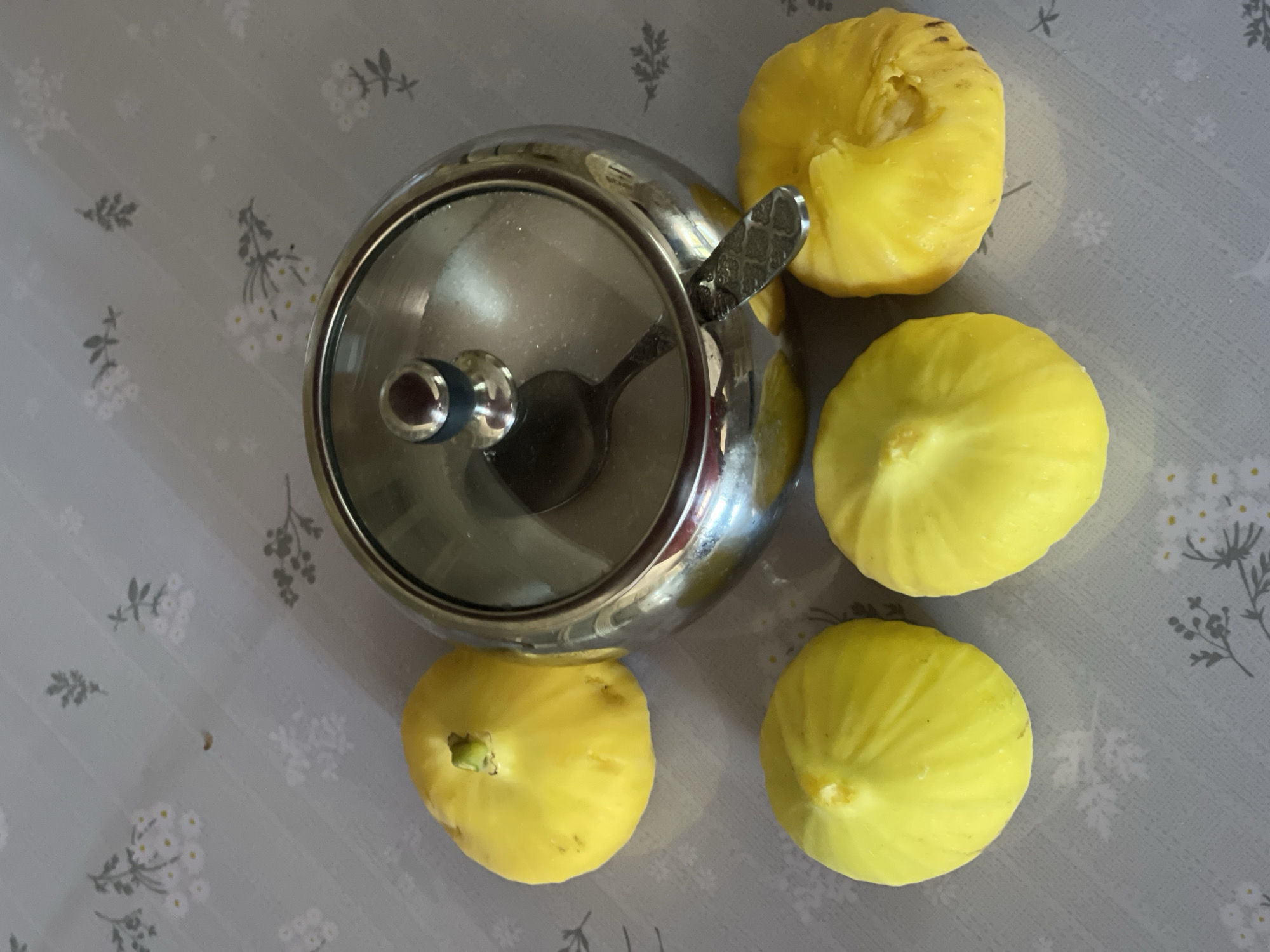 The image depicts a still life arrangement of five yellow figs and a metallic sugar bowl with a glass lid on a grey tablecloth with a subtle floral pattern. The figs are round and plump, with visible stem remnants on some. The sugar bowl is round and shiny, with a spoon resting inside. The tablecloth has a repeating pattern of small white flowers and grey sprigs. The lighting is even, suggesting an indoor setting. The location provided, Tasbuget, Kazakhstan, is not visually evident from the image itself.