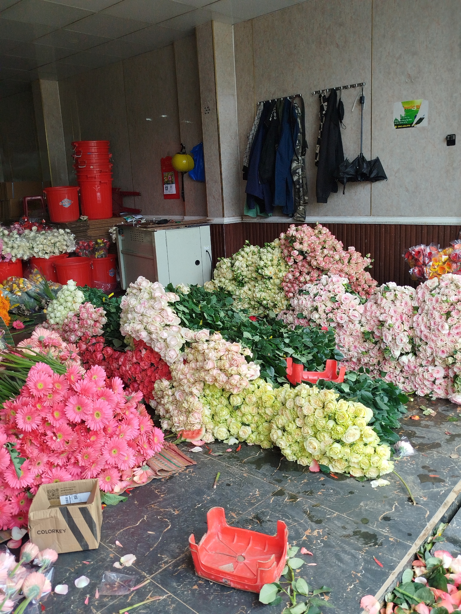 The image captures a flower market scene, possibly in the Cam Ly area of Đà Lạt, Vietnam, as flower cultivation is prevalent in the region. Heaps of various flower types, primarily roses and gerbera daisies in shades of pink, white, and yellow, dominate the foreground and midground. Some flowers are arranged in piles, while others are scattered on the floor, suggesting recent handling or display. Red plastic crates are visible, possibly used for transport or display. In the background, red buckets are stacked near a structure that could be a small refrigerator or storage unit. Clothing items and an umbrella hang on a rack mounted on the wall, suggesting a covered or indoor market space. The floor is tiled and shows signs of flower debris and water, indicating the market is in operation.