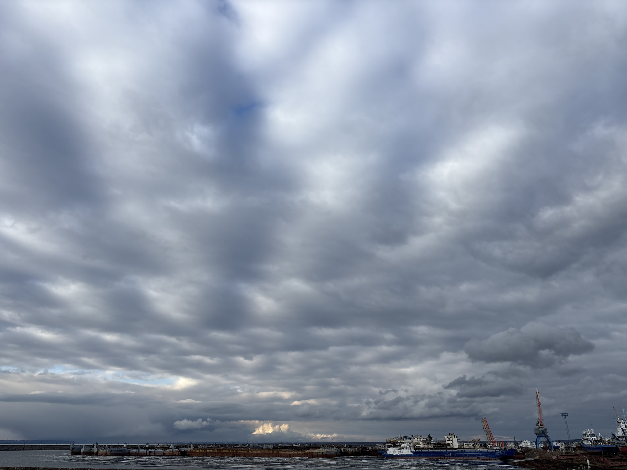 An outdoor, wide-angle shot captures a vast, heavily clouded sky above a large body of water, likely a river or reservoir, with an industrial port in the midground. The sky is dominated by layers of dark grey and lighter grey stratocumulus clouds, with some brighter patches where light penetrates or reflects, suggesting an overcast day.
In the lower portion of the image, the dark water surface shows numerous floating chunks of ice or debris, indicating cold temperatures. Along the distant shoreline, a series of large vessels, including cargo ships and barges of varying sizes and conditions (some blue and white, others rusty), are moored. These ships stretch across the horizon. Prominently on the right, two tall, industrial cranes, one red and white, and another blue and white, rise above the docked ships, characteristic of a working port or shipyard. Further structures, possibly buildings or docks, are visible behind the vessels.
No people are discernibly visible, and no specific activity like loading or unloading is clearly taking place, though the scene suggests an active industrial port. The time of day appears to be daytime, under gloomy, overcast conditions. There is no visible text in the image. The setting, given the context, is likely a port along the Kama River or Votkinsk Reservoir in Chaikovsky, Russia. The overall mood is one of industrial calm under a cloudy sky.