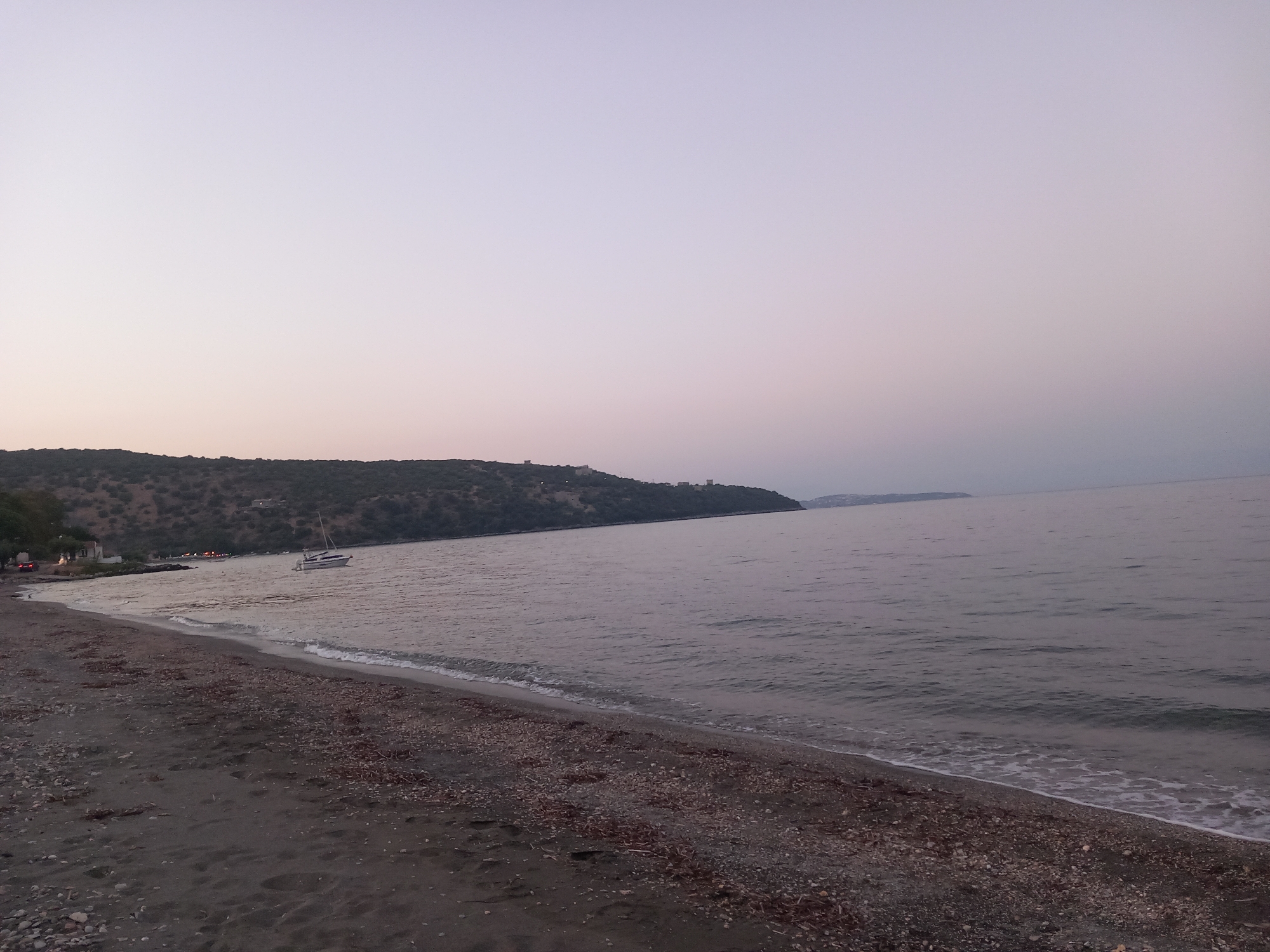 A wide shot captures a serene beach scene at dusk in Gytheio Municipal Unit, Greece. The foreground is dominated by a dark, pebbly beach, with scattered organic debris. A gentle tide laps at the shore, creating a thin line of white foam. In the middle ground, the calm, gray sea stretches towards a distant, hilly coastline. A white sailboat with a tan sail is anchored offshore, its mast a thin vertical line against the muted sky. The sky itself is a soft gradient of pale purple and pink, indicating the transition from day to night. Along the distant shoreline, hints of structures and trees are visible, suggesting a settlement nestled into the landscape. The overall atmosphere is peaceful and tranquil, with no visible people or immediate activity.