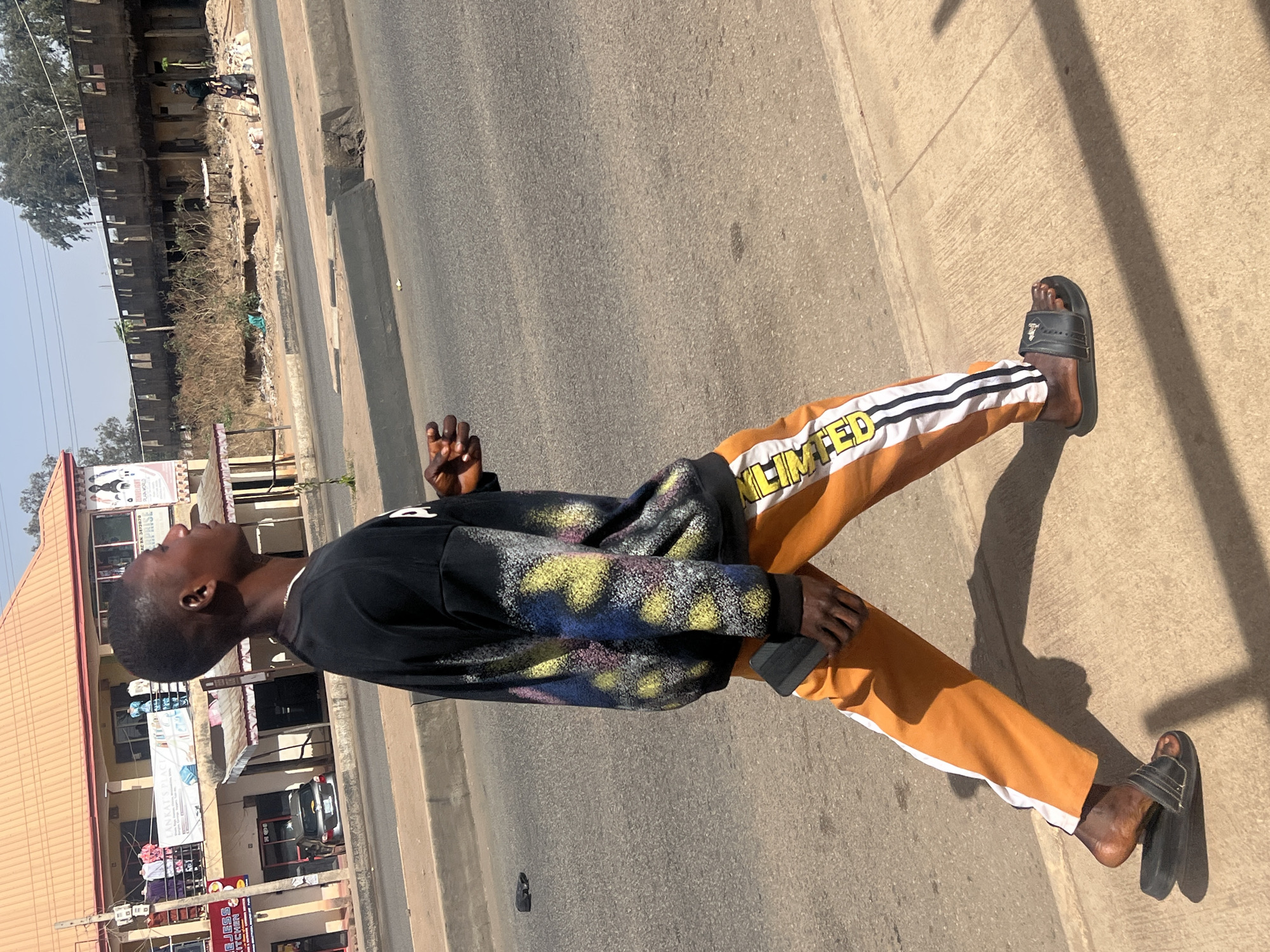 The image depicts a young Black man standing on a concrete sidewalk next to an asphalt road. He is positioned slightly off-center, gazing upwards and to his left. He is dressed in a black long-sleeved sweatshirt adorned with abstract, splattered patterns in yellow, blue, and hints of pink or purple on the sleeves and front. He wears orange pants featuring white stripes along the outer seams, with the word "LIMITED" visible in yellow lettering on the right leg. His footwear consists of black slide sandals. His left hand is raised, appearing to hold a dark, rectangular object, possibly a phone.
The scene is bathed in bright sunlight, casting prominent, dark shadows on the sidewalk and road, suggesting it is midday or early afternoon. The sky above is a clear, light blue.
On the far side of the road, there is a wide, dusty expanse with sparse, dry vegetation and scattered refuse. Beyond this area stands a row of multi-story buildings; some appear to be unfinished concrete structures with visible scaffolding or empty window frames, while others are more developed commercial buildings with corrugated metal roofs and various storefronts. Trees are visible further in the background.
Behind the man, on the same side of the road, are several commercial buildings with visible signs and awnings. Some legible text includes "ENTERPRISE" on a sign above a storefront and "KITCHEN" on another. A dark-colored car is visible on the road near these buildings. The overall setting portrays a typical urban or semi-urban street environment in Jos, Nigeria.