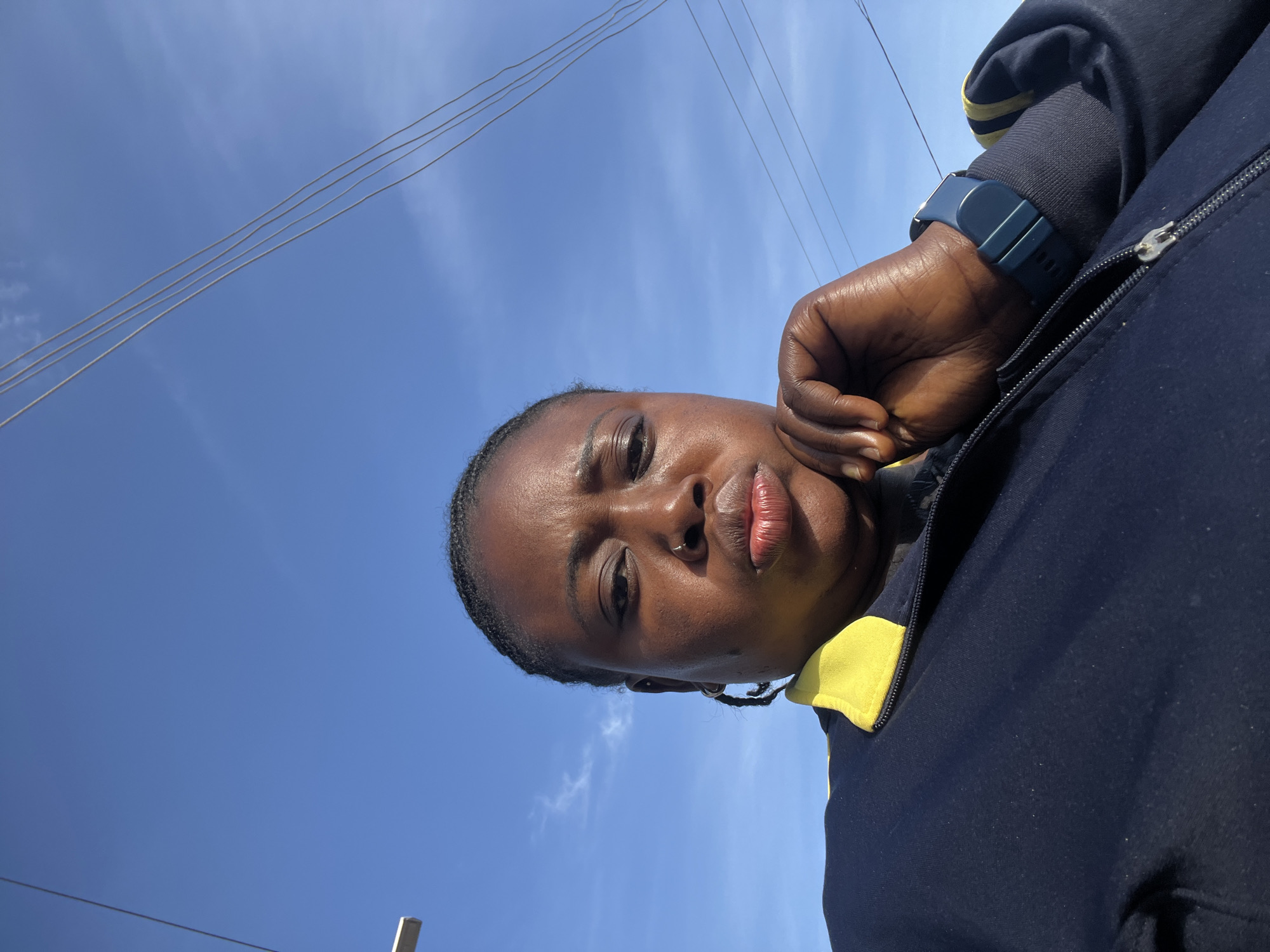 A low-angle, close-up, selfie-style photograph shows a Black woman from the chest up, looking at the camera with her chin resting on her left hand. She has her lips pursed and wears a navy blue jacket with a yellow collar. Her hair is braided into cornrows, and she has a nose ring. A blue smartwatch is visible on her left wrist. The background is a clear blue sky with a few wispy clouds and power lines. The scene is outdoors, likely in the city of Jos, Nigeria, given the style of power lines and the presence of a street light visible on the left edge of the frame. The lighting suggests it is daytime.
