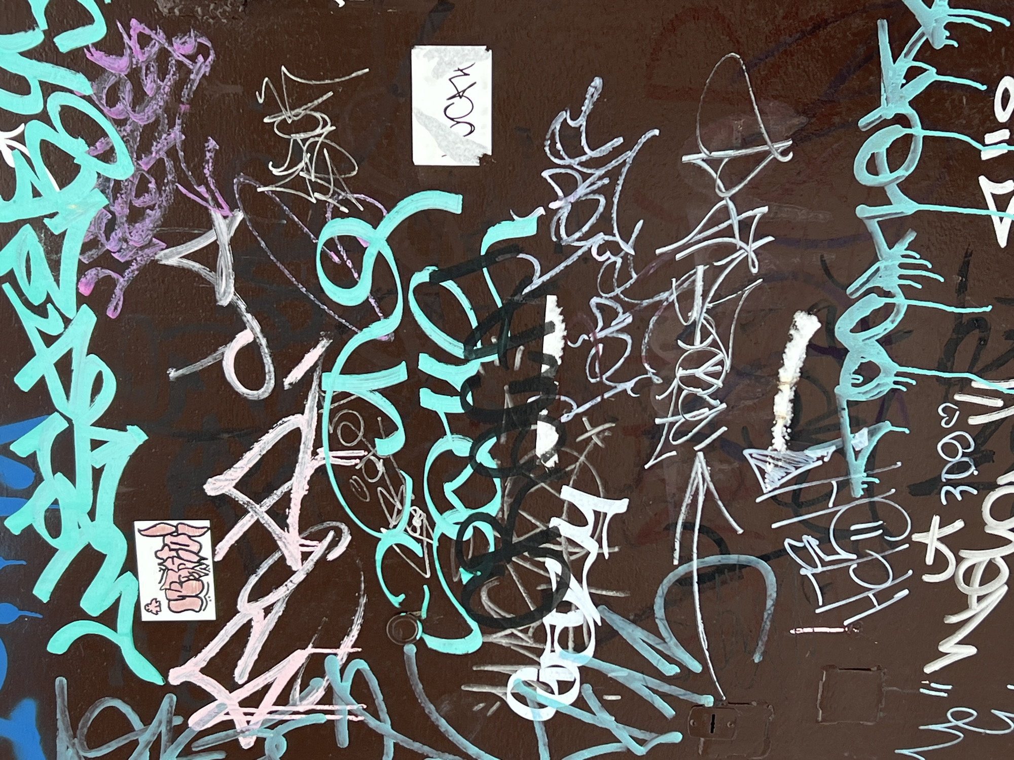 The image depicts a densely tagged and stickered dark brown surface, possibly a wall or large metal object. A variety of graffiti styles are present, using a range of colors including bright teal, white, black, and pale pink. The graffiti consists primarily of overlapping tags and abstract line work, with some instances of legible letters and words. Two stickers are adhered to the surface: one with an illegible stylized design in pink and white, and another small white sticker with handwritten text. The lower right corner reveals a small keyhole and lock mechanism. The location of this image is Chaikovsky, Russia.