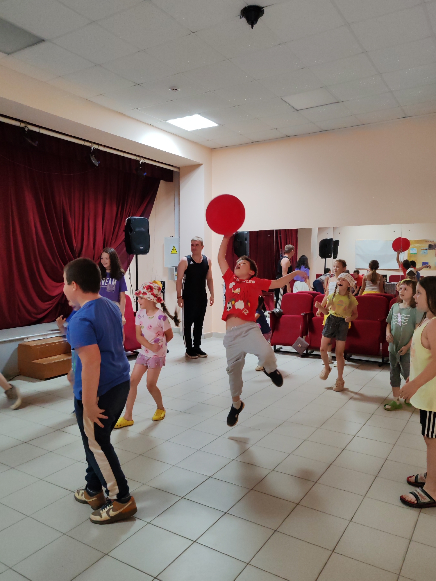 In a room with light-colored tiled flooring and pale peach walls, a group of children and at least one adult are engaged in an activity. A red balloon is held aloft by a young boy in a red t-shirt and grey pants, who is in mid-air, jumping. Other children are scattered throughout the room, some observing, others participating. A teenage girl in a purple t-shirt stands near a red velvet curtain and a speaker. A man in a black tank top and black pants is positioned to the right of the curtain. The environment suggests a recreational or event space, possibly a community center or a hall, with a stage area partially visible behind the red curtain. The room also contains red chairs, mirrors, and a white board. The location is identified as the city of Чухлэм, Russia.