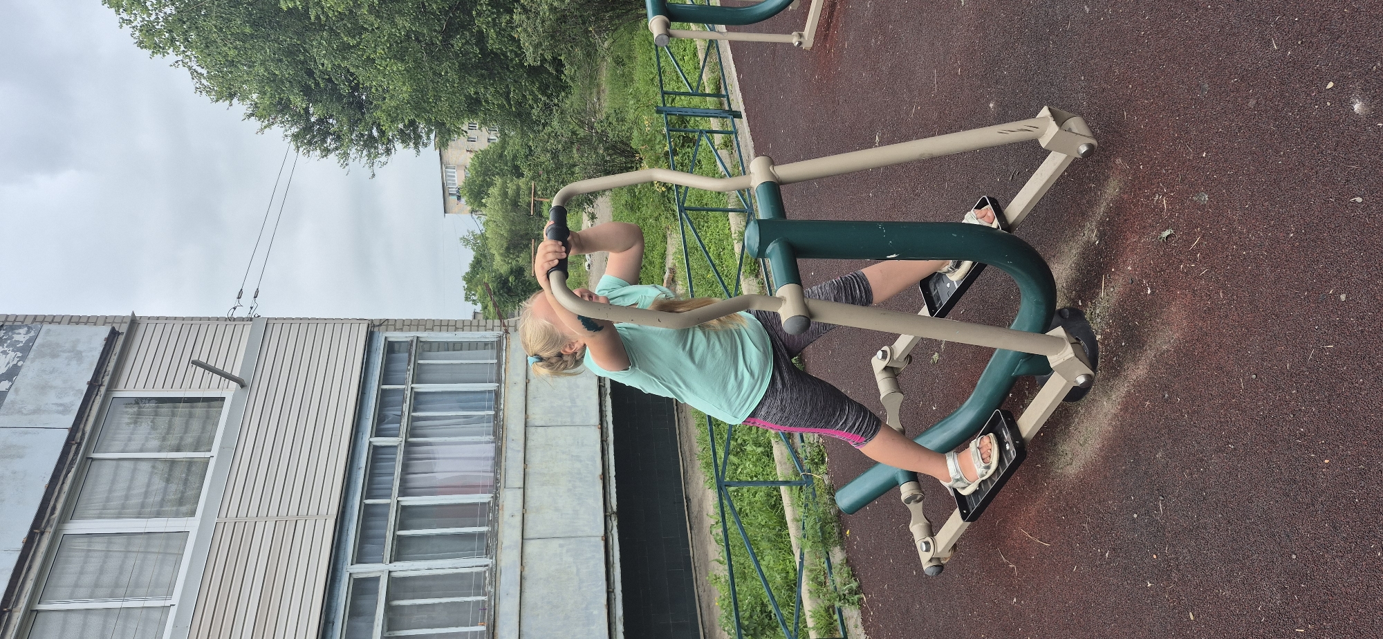 A blonde girl with her hair in a ponytail is exercising on an elliptical machine outdoors. She is wearing a light blue t-shirt and grey capri pants with pink stripes. She is holding onto the handles of the machine and appears to be in motion. The elliptical machine is green and beige. The ground is covered in a dark red rubberized surface. In the background, there is a building with balconies and windows, a green metal fence, and trees. The sky is overcast.