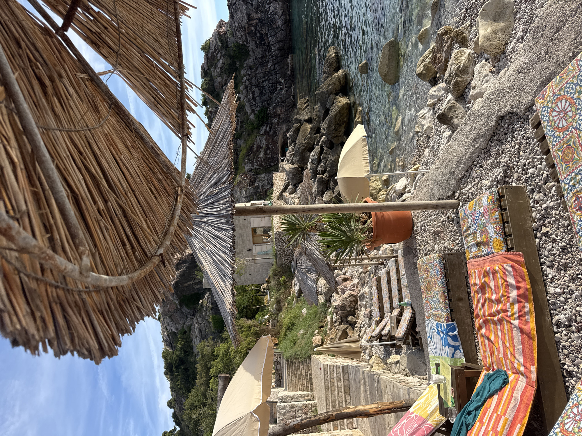 The image depicts a tranquil beach scene in Rijeka Reževići, Montenegro. The foreground features several wooden loungers, each adorned with vibrantly colored towels and patterned cushions, suggesting a comfortable and inviting atmosphere. Behind the loungers, a pebble beach slopes down to the clear, calm water of the Adriatic Sea, which is dotted with large rocks. The mid-ground reveals more beach chairs and a few straw umbrellas providing shade. A stone pathway with steps ascends the hillside, leading towards a small, rustic stone building. Lush green vegetation and rocky outcrops characterize the rugged coastline in the background. The sky is a clear blue with a few wispy clouds, and the sunlight casts long shadows, indicating it is late afternoon or early evening. No people are visible in the frame.