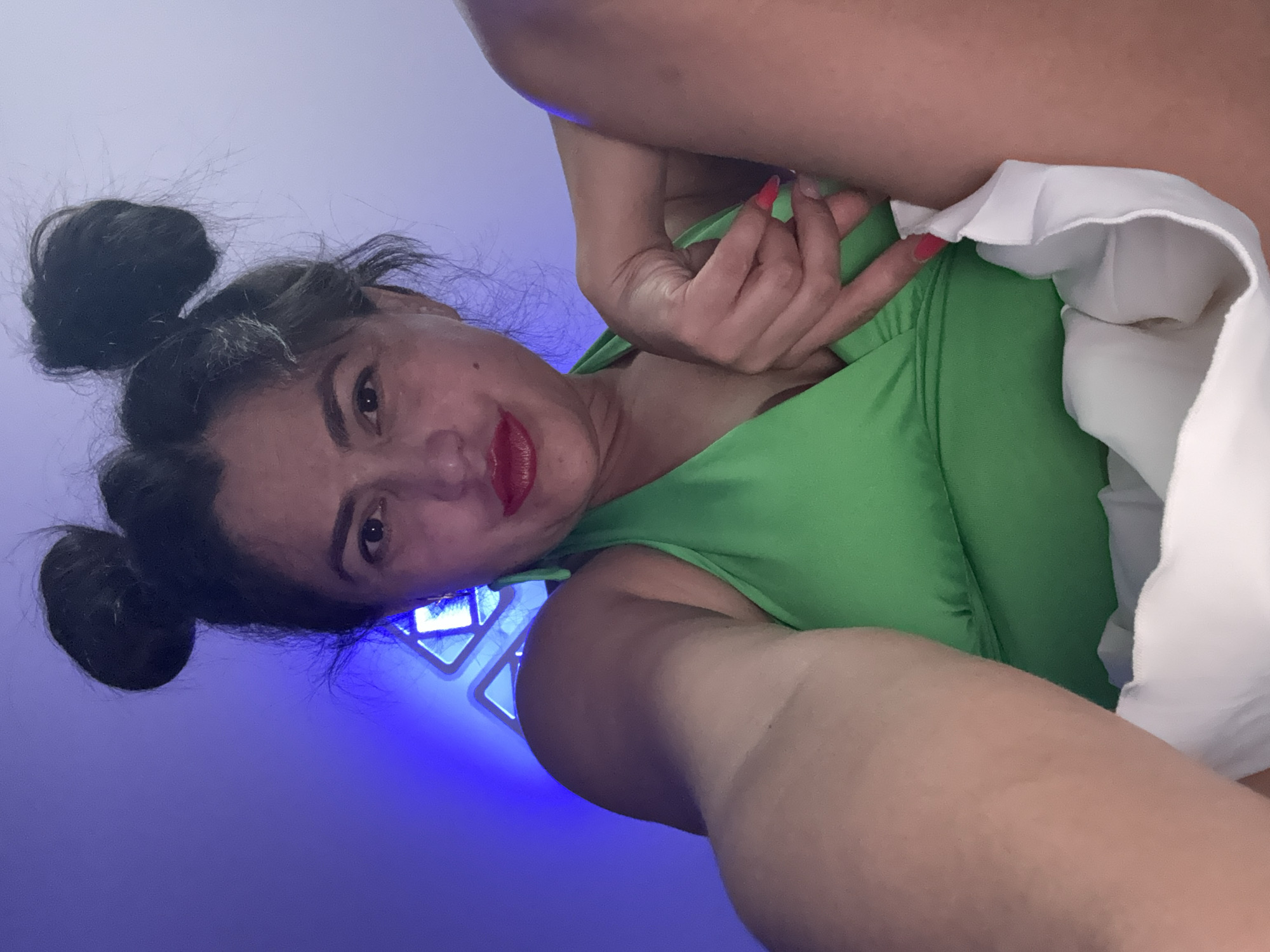 The image presents a close-up portrait of a woman, oriented sideways, captured from a low angle. She has dark hair styled into two distinct buns on the top of her head. Her face is turned towards the viewer, featuring dark eyes, well-defined eyebrows, and vibrant red lipstick, with a slight smile or smirk. She is dressed in a bright, almost neon green top or dress. Her right hand, with fingers painted with bright red nail polish, is visible near her chest, appearing to adjust her attire or hair. A white garment with soft ruffles is also visible on her right side. The background is dominated by an intense blue light, possibly from LED panels or a colored light source, creating a strong ambient glow. Geometric shapes, likely light fixtures, are visible behind her head and shoulder, emitting this blue illumination. The setting appears to be indoors, likely at night or in a dimly lit space, with artificial blue light serving as the primary light source. The woman's direct gaze suggests a posed photograph or a selfie. The location context for this image is Kauchuk Stantsiya, Russia.