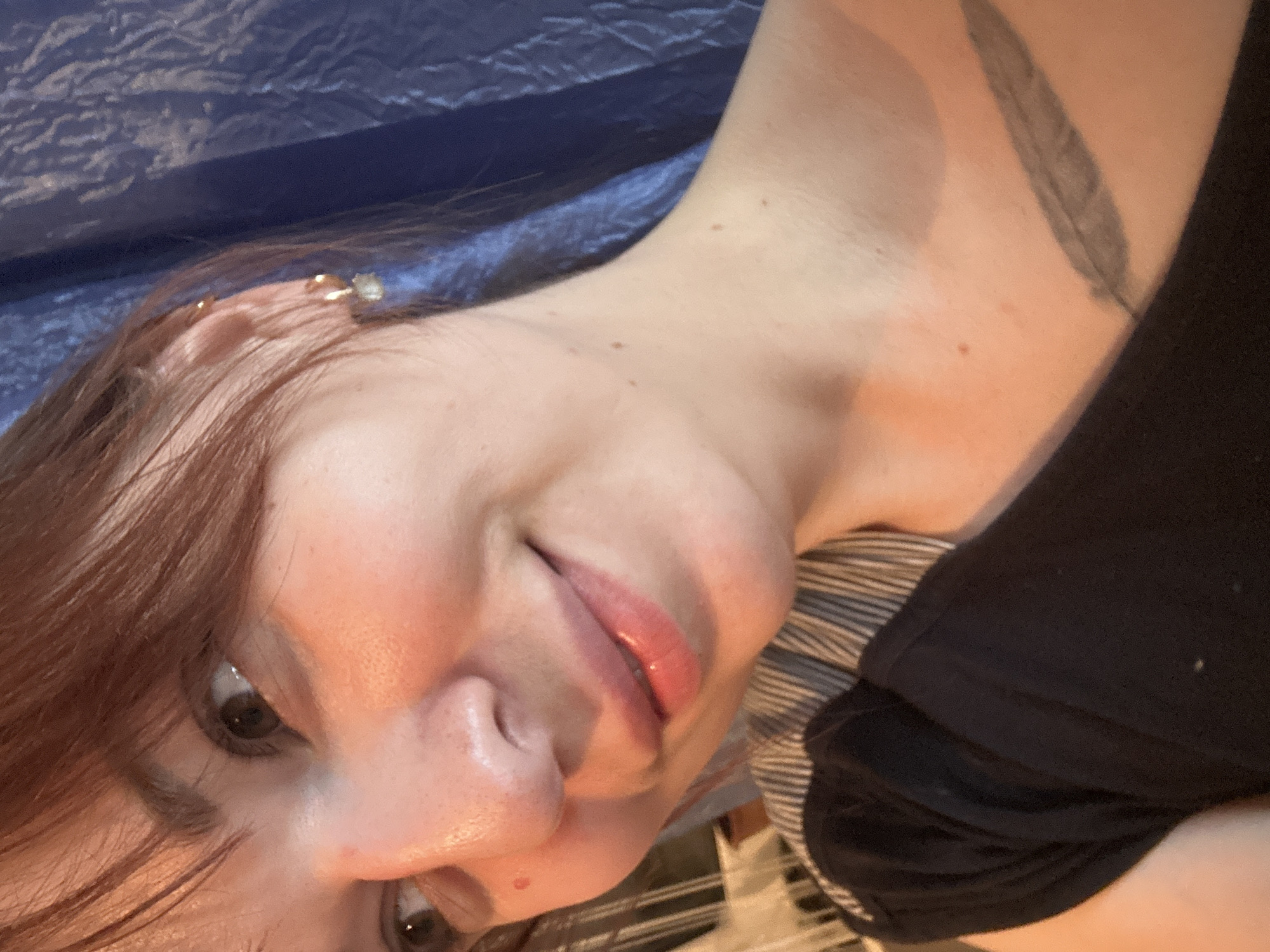 A selfie shows a woman with fair skin and auburn hair. She has brown eyes, and her lips are slightly parted. She has an earring in her upper ear. A tattoo resembling a feather is visible on her upper shoulder/neck area. She is wearing a black shirt. The background is a blurry blue, possibly a fabric or surface. There is no indication in the image of the location being in Фоки, Russia.