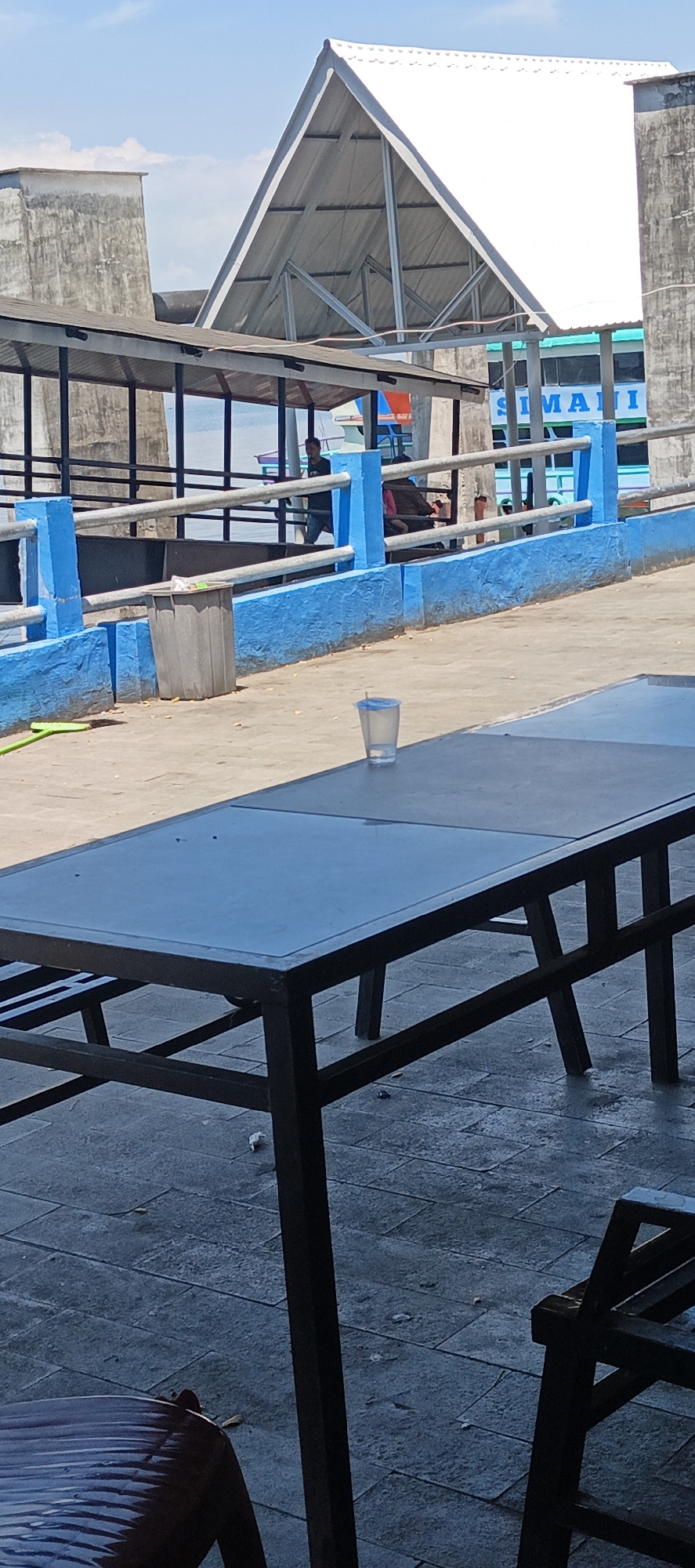 The image shows a scene at what appears to be a ferry terminal or dock in Tigaras, Indonesia, on a sunny day. In the foreground, a dark-colored rectangular table with a reflective surface is positioned on a patterned concrete ground. To the left of the table, the edge of a dark chair with a textured seat is visible. On the table, a clear plastic cup containing a clear liquid and a blue straw is present.
Behind the table, a walkway with a bright blue painted wall and a grey metal railing runs along the left side of the frame. A grey trash can with some items inside is located near the blue wall. Further back, a covered walkway with a dark roof supported by dark poles extends into the scene. Through the railings and the covered walkway, a body of water and a boat with the name "SIMAHI" visible on its side can be seen. Several people are present in the middle ground, some standing and others seated near the railing. One person is visible under the covered walkway, and another appears to be walking.
The background features a large, light-colored triangular roof structure supported by metal beams, and a tall, grey concrete wall on the far left. The sky is blue with scattered white clouds. The overall environment suggests an outdoor public space in a waterfront location.