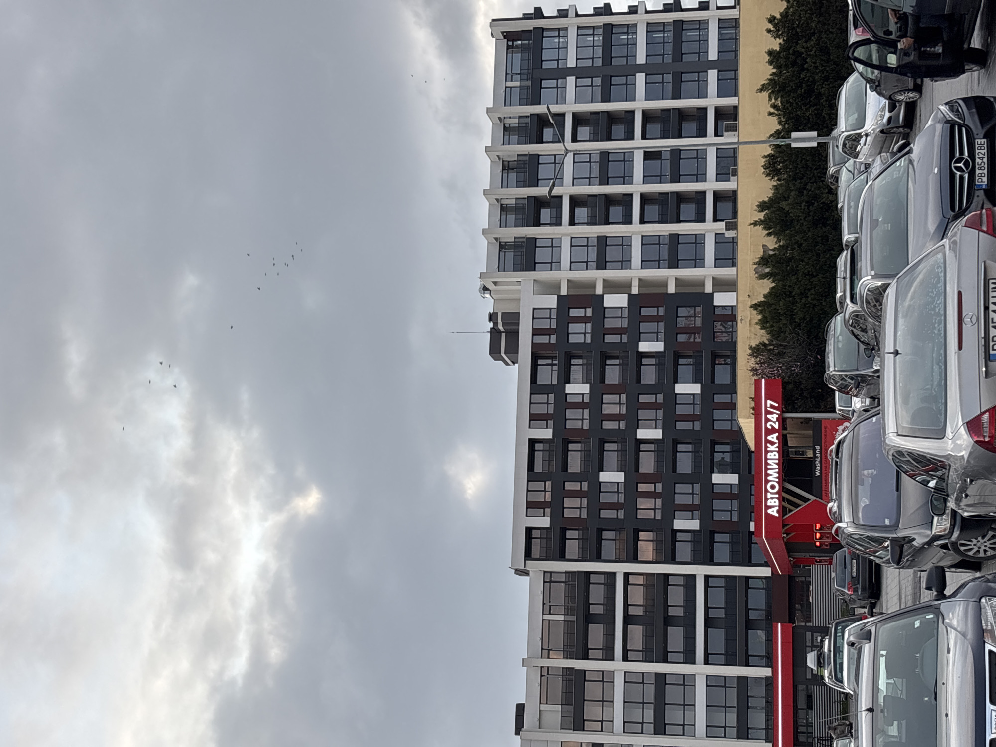 The image shows a modern high-rise building with a black and white facade in Plovdiv, Bulgaria. Next to the building is a 24/7 car wash ("АВТОМИВКА 24/7" in Cyrillic). Several parked cars are visible in front of the building and the car wash. The sky is overcast with cloudy. Several birds are flying in the sky.
