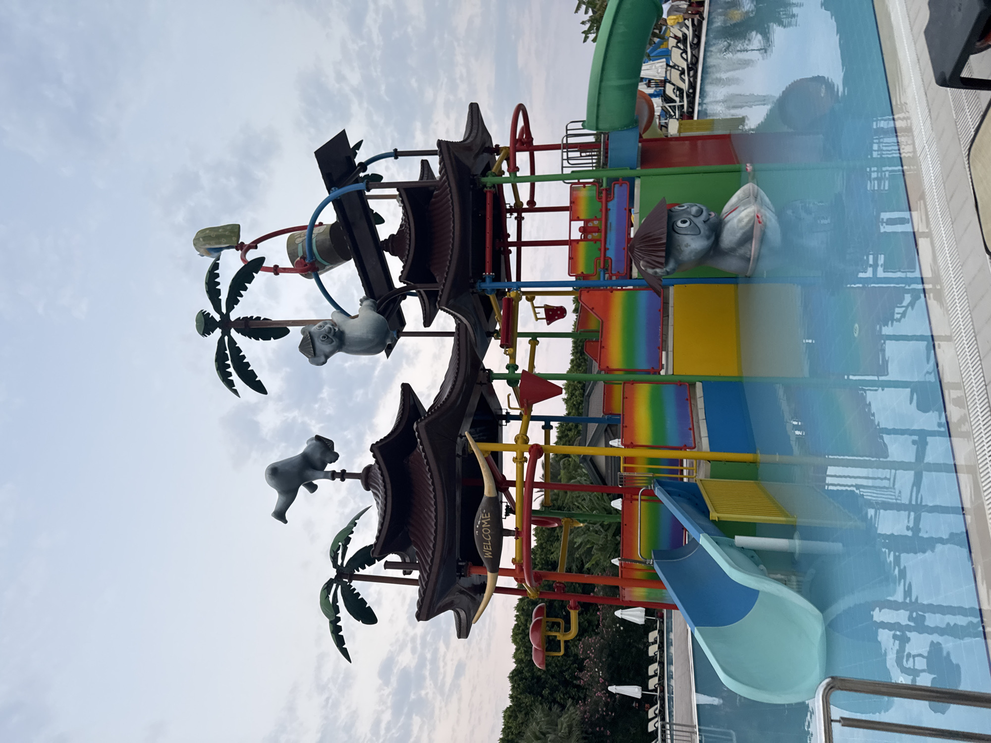 This image depicts a brightly colored water park attraction with a central pagoda-style structure. The structure has multiple levels and is adorned with cartoonish animal figures, including a monkey-like character on top and a koala-like figure near a bucket. Palm tree decorations are also visible. Various slides, including a blue and green slide, are integrated into the attraction, all situated within a clear blue swimming pool. The pool area is surrounded by lounge chairs and umbrellas, suggesting it is part of a resort or hotel. The sky is overcast. The location is identified as Aksu, Turkey.