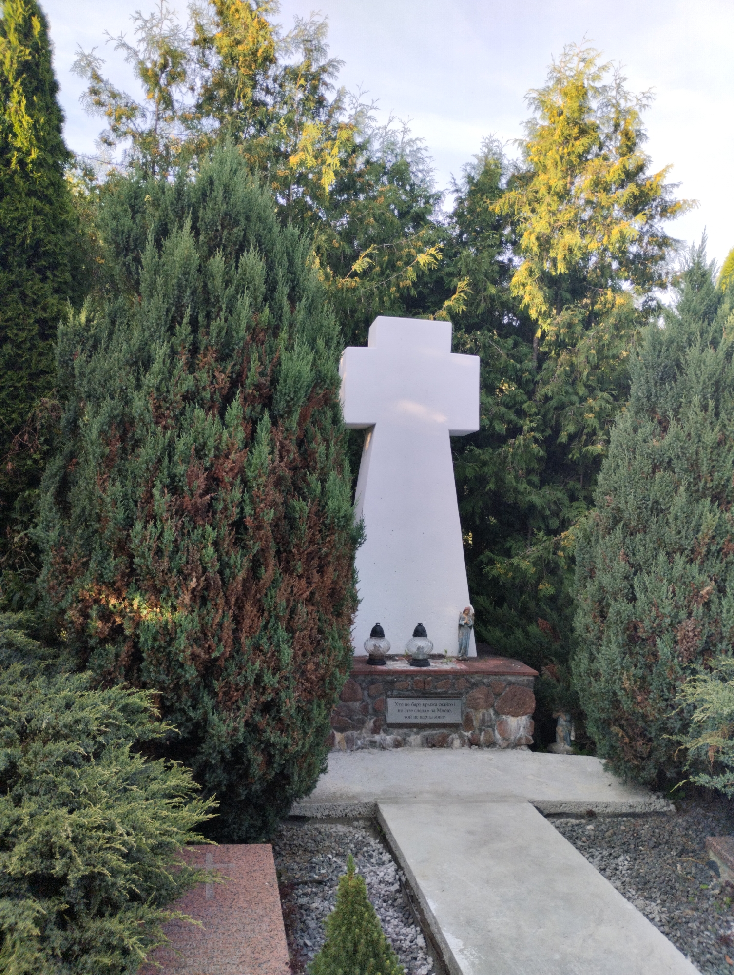 A wide, outdoor, eye-level shot depicts a white cross grave marker in the middle of a cemetery. The cross is tall and angular, with a rounded base. In front of the cross, there is a low, stone wall surrounding the base. On top of the wall, there are two glass grave lanterns with black lids and a small angel figurine. A stone plaque is affixed to the wall. Surrounding the grave marker are mature evergreen trees and bushes. A concrete pathway leads to the grave. The ground is covered with gravel and a red granite tombstone is visible on the left side of the frame. The sky is clear and bright, indicating daytime. The location is identified as the cemetery in the village of Vishnevsky, within the Vishnevsky rural council, Belarus.