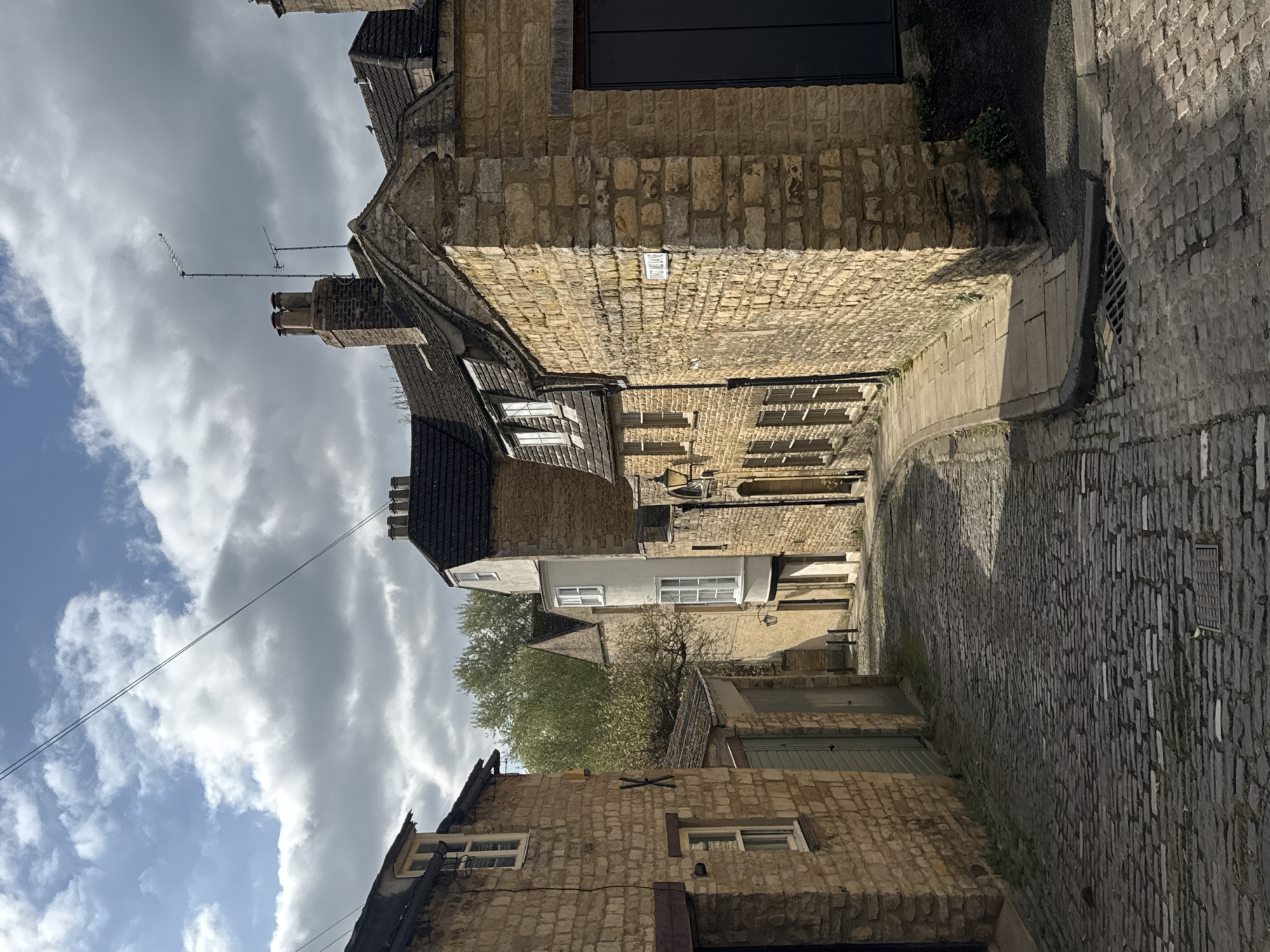 This vertical image depicts a narrow, historic alleyway or street winding between traditional stone buildings under a partly cloudy sky. The perspective looks down the cobbled path as it curves gently to the right, leading deeper into the architectural setting.
The buildings on both sides are constructed from light-colored, rough-hewn stone, characteristic of historic British architecture, consistent with the South Kesteven location. Many structures feature dark, possibly slate or tiled, roofs with prominent brick chimneys and chimney pots. Various windows, some with distinct white frames, are visible across the stone facades.
The ground is paved with well-worn cobblestones, indicating an old and well-trodden pathway. Shadows cast by the buildings suggest bright daylight, likely late morning or early afternoon, with sunlight illuminating parts of the street and buildings, while other areas remain in shadow.
Notable details include multiple television aerials or antennas mounted on rooftops, silhouetted against the sky. Power lines or cables stretch across the scene, connecting between poles or buildings. A small white rectangular street sign is visible on the stone wall of a building on the left, but the text on it is largely unreadable. Patches of greenery, possibly ivy or small bushes, sprout from crevices in the stone walls and along the street's edge. No people are present, contributing to a quiet, serene, and timeless atmosphere.