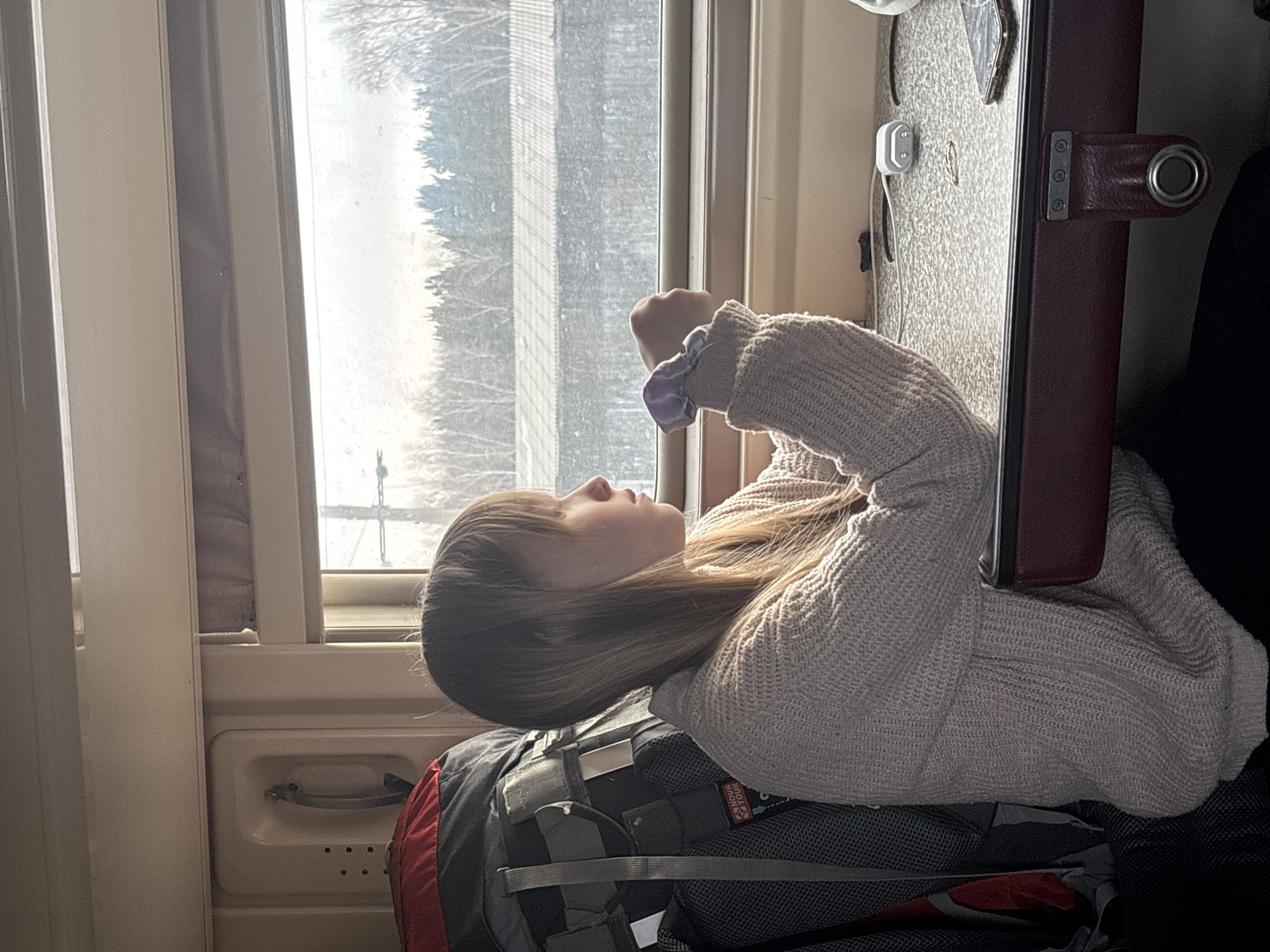 A young female is depicted seated inside a train compartment, gazing out a window at a landscape consistent with travel through the vicinity of Usolye-Sibirskoye, Russia. The subject, appearing to be a young adult, has long, straight, light brown hair and is wearing a light beige, chunky knit sweater over dark trousers. A light purple scrunchie is visible on her left wrist. Her left arm rests on a light grey, speckled table with a dark red upholstered edge, where a white Apple AirPods charging case, a white charging cable, a dark-colored smartphone with a transparent or glitter-flecked case, and a small metallic ring are present. To the left of the female, a large dark grey and red hiking backpack, bearing a "SNOVA TOUR" label, occupies a seat or storage space. The train's interior features light-colored paneling and a cream-colored fixture with a receiver-like handle and perforations. The exterior view through the window shows a pale, overcast sky above a wintery or early spring landscape. This landscape includes bare deciduous trees in the foreground, darker evergreen trees behind them, and a low-lying building with a corrugated roof in the mid-distance. A utility pole with crossbars is partially visible on the left side of the window frame. Natural light from the window illuminates the scene.