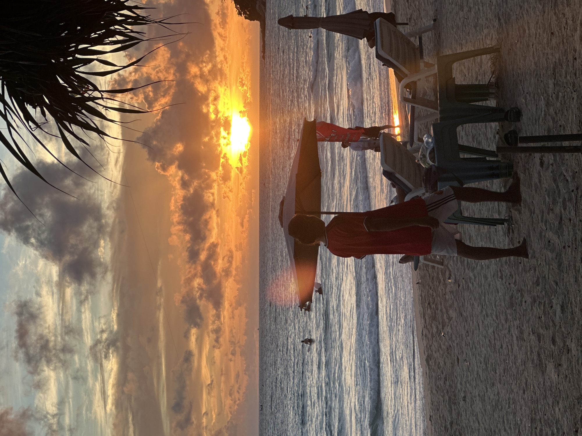 This image depicts a serene beach scene at sunset in Karon, Thailand. The sky is ablaze with vibrant orange and yellow hues, indicating a beautiful sunset. Wispy clouds are scattered across the sky, catching the golden light. In the foreground, palm fronds cast a shadow, framing the view.
The beach is lined with empty lounge chairs and umbrellas, suggesting a quiet time of day or perhaps the end of the day's activities. The calm ocean reflects the warm colors of the sunset, with gentle waves lapping at the shore.
A few people are visible. In the mid-ground, a person in a red shirt and white shorts stands on the sand near the water's edge, facing away from the camera. Further out in the ocean, several small figures can be seen, possibly enjoying a swim or a paddle. The overall mood is tranquil and picturesque, capturing the beauty of a tropical sunset. The sand appears soft and golden, characteristic of a beach setting.