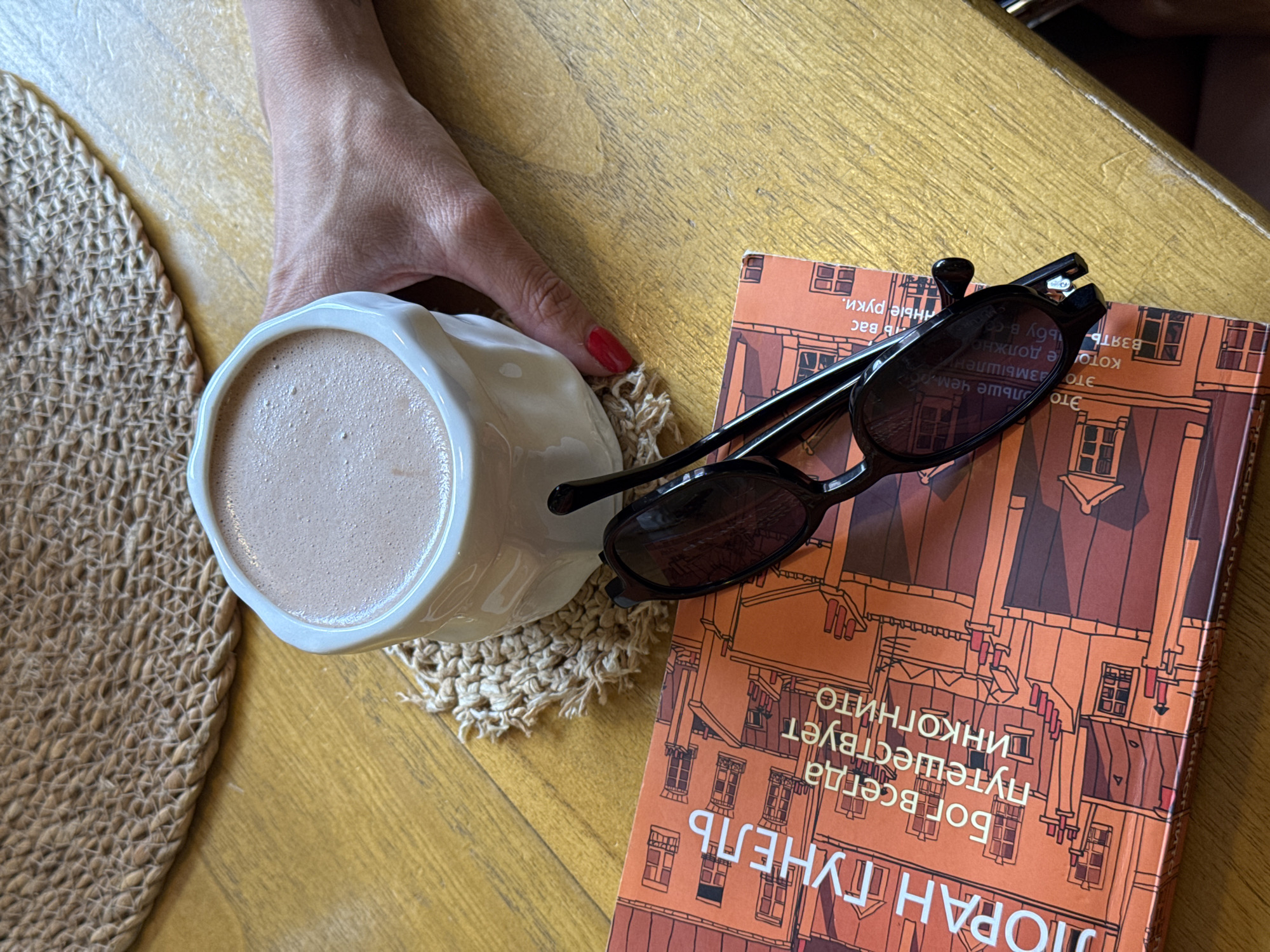 A close-up overhead shot shows a light brown frothy drink in a white mug, placed on a crocheted coaster. Next to the mug is a book with an orange cover, featuring an illustration of buildings and text in a foreign language. A pair of black sunglasses rests on top of the book. A person's hand, with red painted fingernails, is visible near the mug, suggesting they are holding it or about to pick it up. The scene is set on a wooden table with a woven placemat in the background. The overall impression is one of leisure and relaxation. The text on the book appears to be in Russian, and the style of the book cover and the general aesthetic could be found in many cafes or restaurants in Batumi, Georgia.