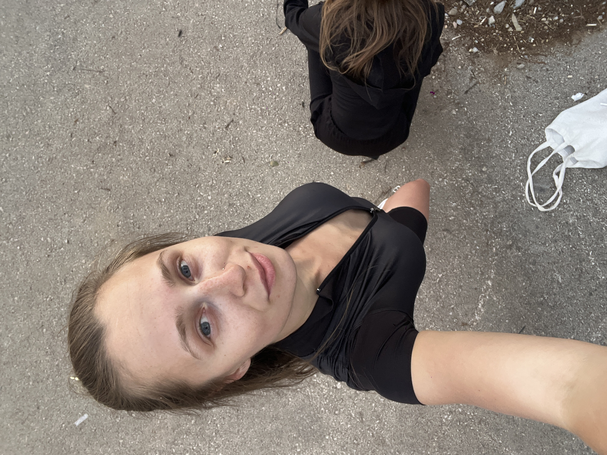 A photograph taken from a high angle shows a young woman with fair skin, light brown hair, and blue eyes, wearing a black fitted shirt. She is holding the camera, as evidenced by her arm visible in the foreground. Behind her, partially visible, is another person sitting on the ground wearing a black hoodie and pants. A white tote bag and a disposable face mask are next to that person. The background is a concrete or asphalt surface, possibly a sidewalk or parking area. In the top right corner, there is some debris. No specific information is available to determine the location within the city of Budva, Montenegro.