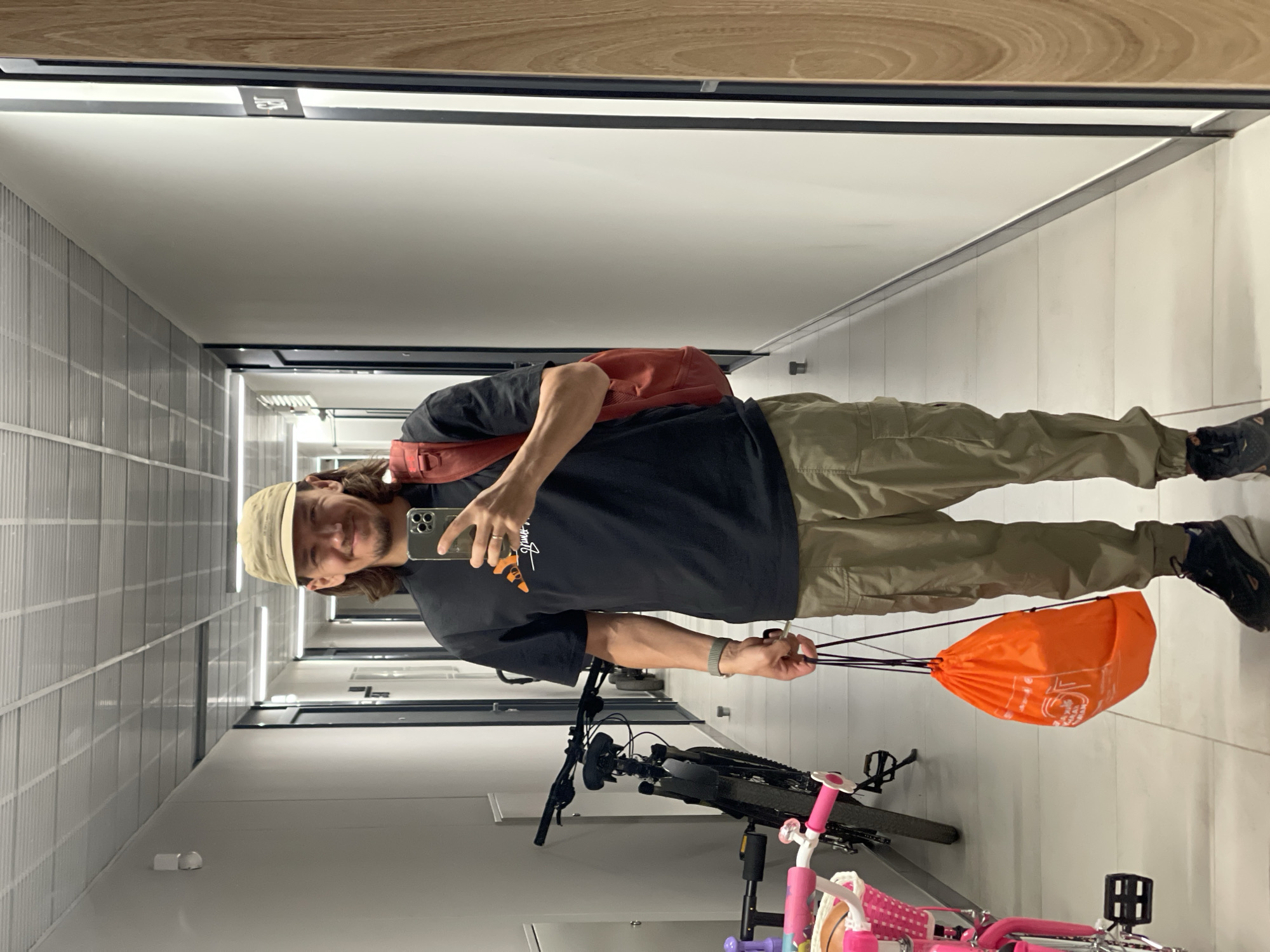 In a dimly lit indoor hallway, a man with medium-length brown hair and a beard stands facing a mirror, taking a selfie with his phone. He is wearing a beige baseball cap, a black t-shirt with an orange graphic, and olive-green cargo pants. A red backpack is slung over his right shoulder, and he holds an orange drawstring bag in his left hand. His right arm is bent, and he wears a watch on his wrist. The hallway features white walls, a tiled floor, and a suspended ceiling with fluorescent lighting. To his left, a black bicycle is parked, and a pink children's bicycle is partially visible in the foreground. The hallway extends into the distance, with multiple doors visible on either side.