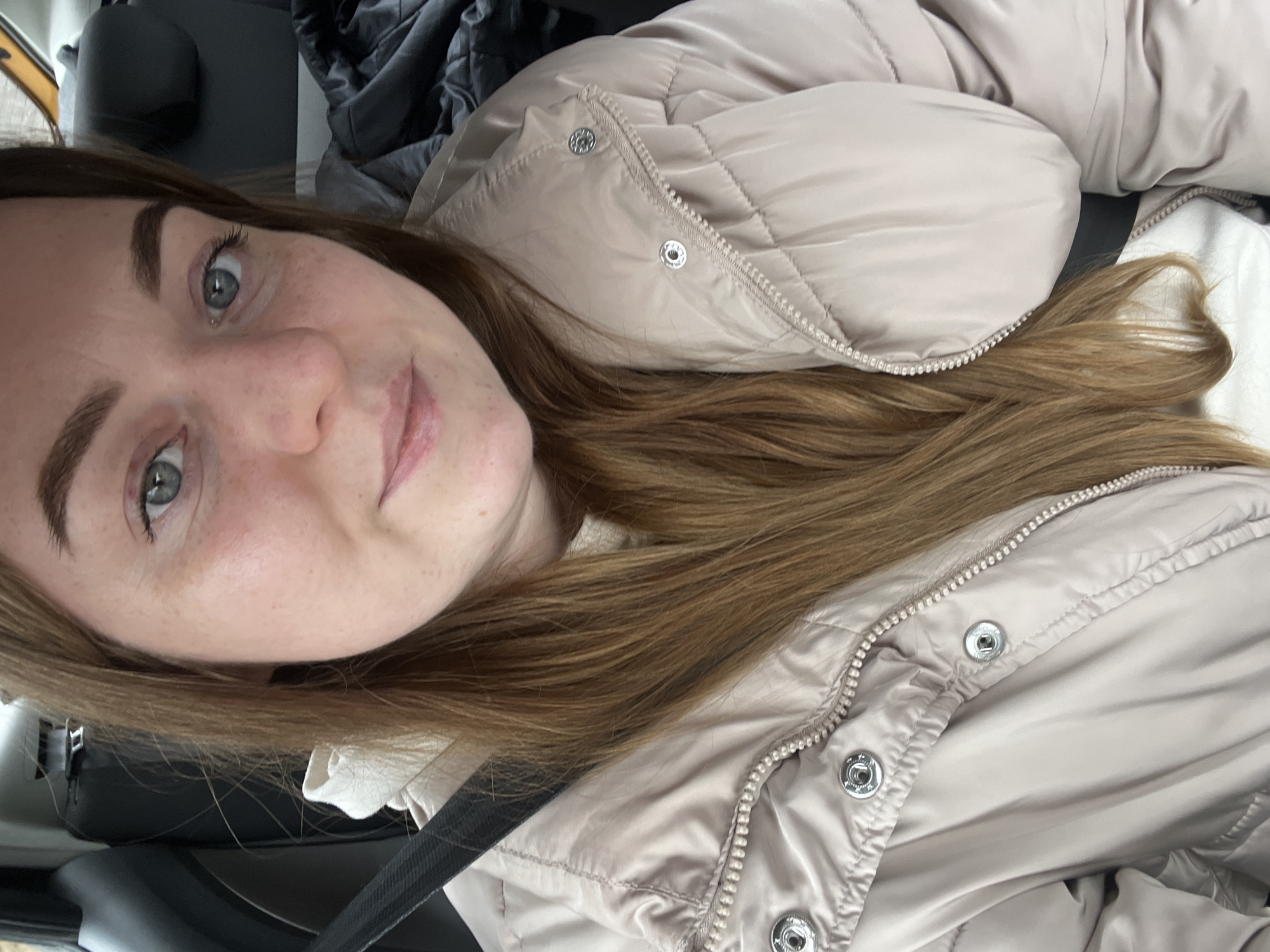 A selfie of a woman with light skin and freckles is taken from a low angle. She has light brown hair and blue eyes. She is wearing a light beige puffer jacket. A car seatbelt is visible across her chest. The background includes the interior of a vehicle, with dark upholstery and other items partially visible. The location in Rogachovo, Russia, cannot be determined from the visual information.