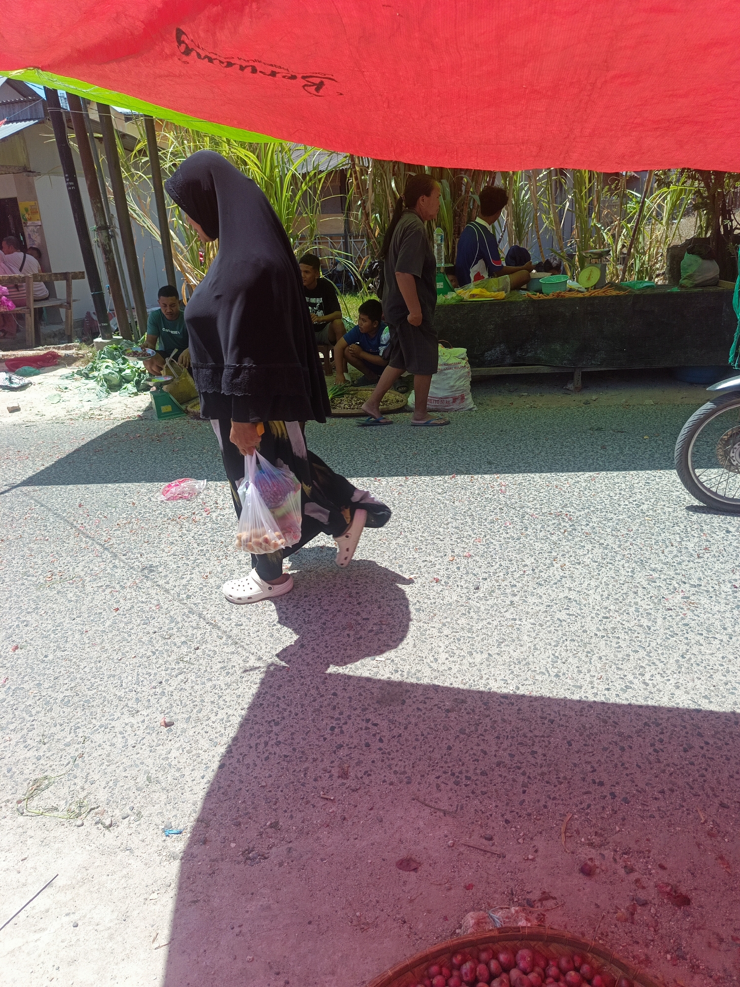 The image depicts an outdoor market scene, likely in Gorontalo, Indonesia, under a large red and green tarp. In the foreground, a person wearing a black hijab and a patterned skirt walks from left to right, carrying a transparent plastic bag filled with various items. White Crocs are worn on their feet. A distinct shadow of this individual is cast on the asphalt ground, which is littered with small red debris.
In the background, under the tarp, several people are visible. One person in a green shirt is seen near a green scale, with what appears to be durian fruit on display. Another individual in a blue and white shirt is standing behind a market stall laden with produce, including what looks like yellow fruits and leafy green vegetables. Further back, a person in a dark t-shirt and shorts is sitting, and a child in a blue shirt is also present. In the far right of the image, the rear wheel of a motorcycle is visible. The environment suggests a sunny day, with tall grass or bamboo visible behind the market stalls. The ground is a mixture of asphalt and sand, with various small objects scattered on it.