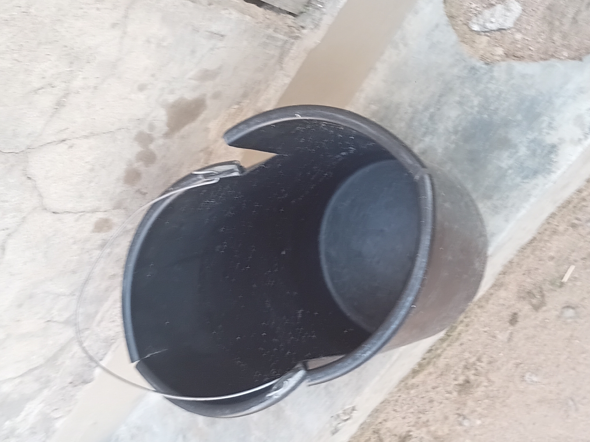 An outdoor, close-up, slightly high-angle shot shows a black plastic bucket with a metal handle. The bucket is positioned on a concrete surface next to a dirt area. The background consists of a textured, light-colored wall with some cracks and stains. The scene appears to be in Jalingo, Nigeria.