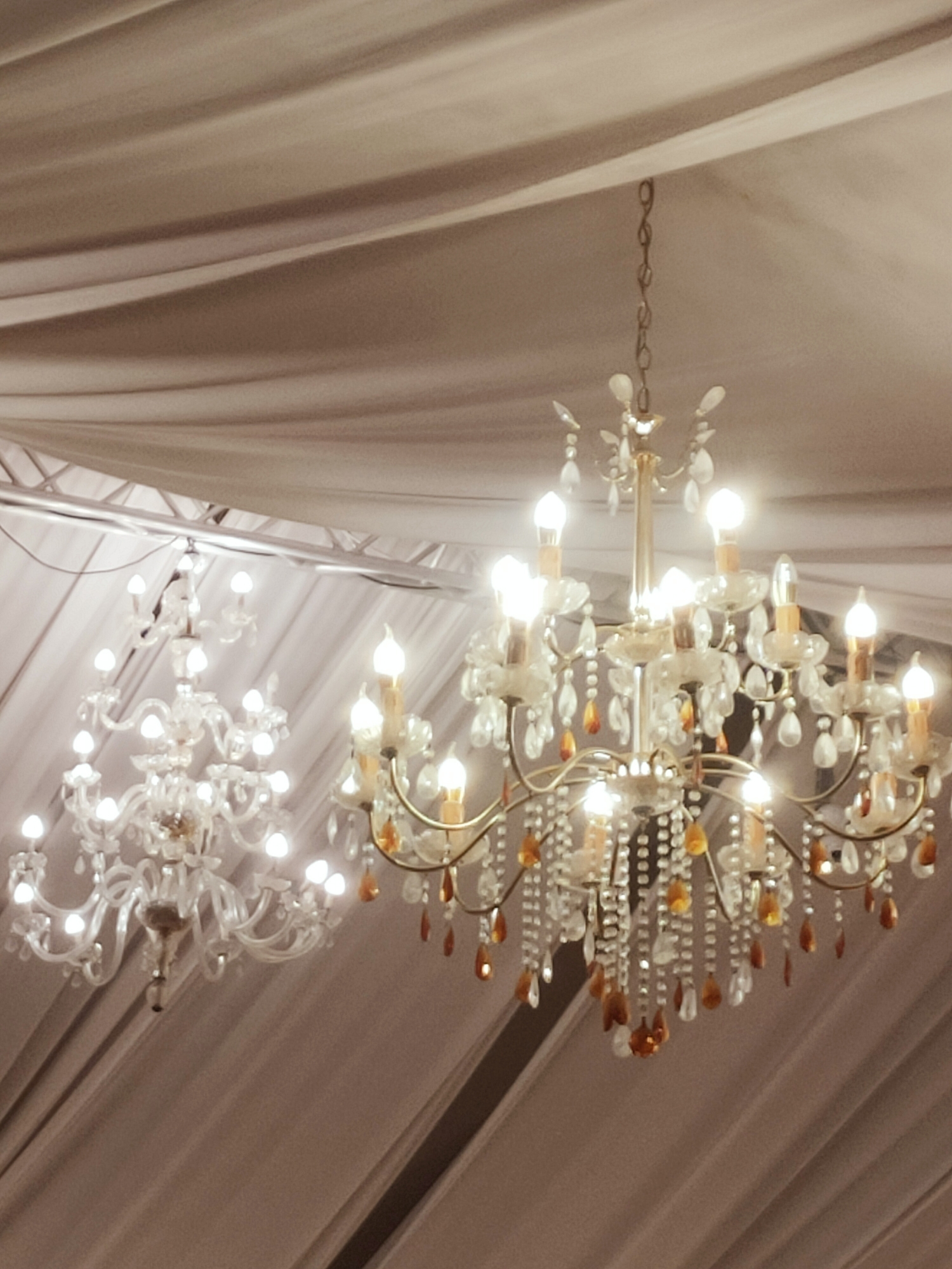 Two ornate chandeliers hang suspended from a draped ceiling in what appears to be a formal event space. The chandelier in the foreground is larger, with a gold-toned metal frame, adorned with numerous clear crystal droplets and amber-colored tear-shaped beads. It features multiple bulbs emitting a warm light. Behind it, a second, smaller chandelier with a clear glass or crystal structure and similar droplet embellishments is partially visible. The ceiling is covered with flowing, light-colored fabric that creates soft folds and shadows. The background is a muted, brownish-grey color. The scene suggests an interior setting, possibly a banquet hall or wedding venue in Mornag, Tunisia. No people are present in the image.