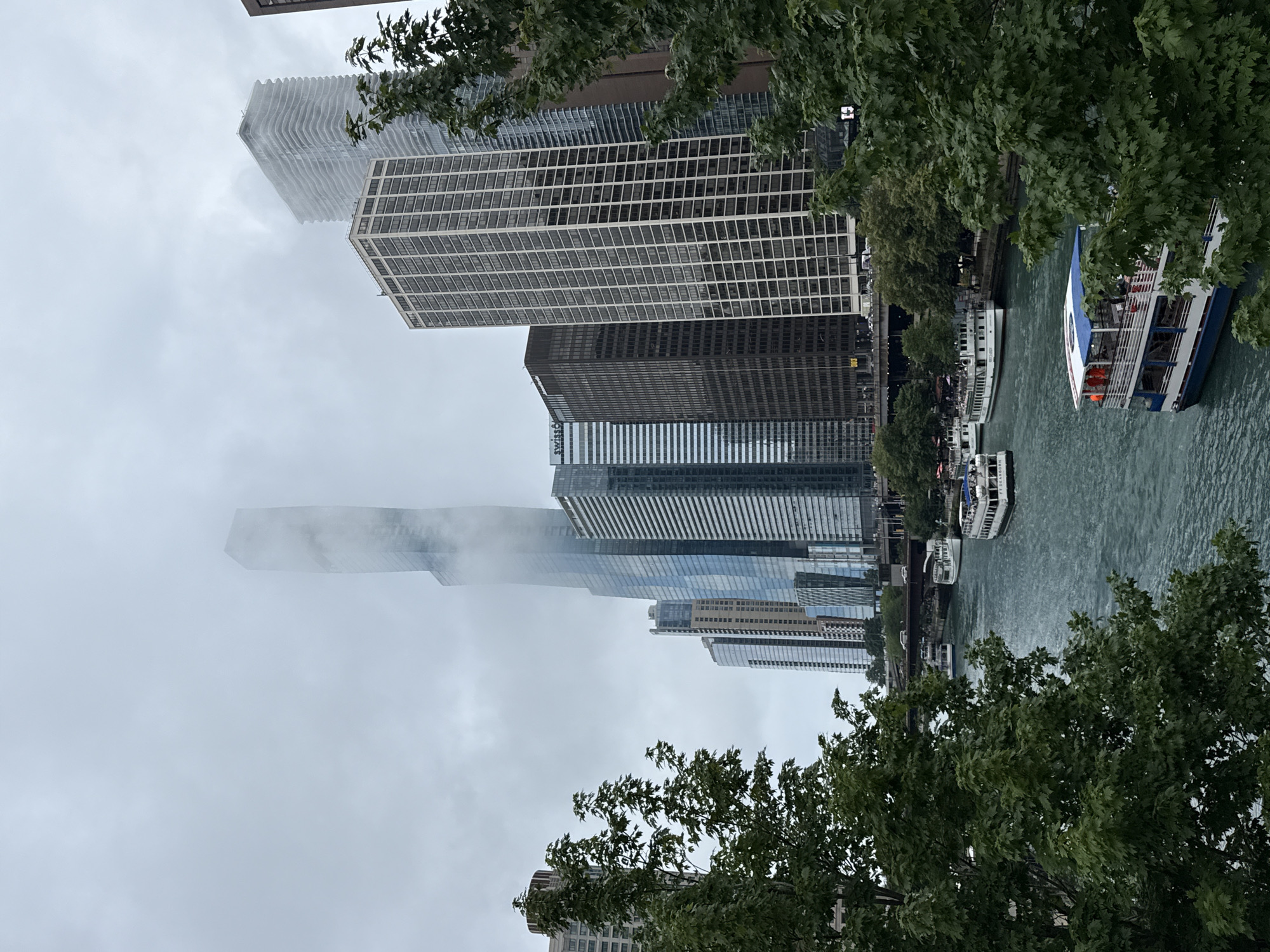 This is a photograph of the Chicago River in Chicago, Illinois, United States of America. The scene depicts a cloudy day with a view of various skyscrapers lining the river. Several tour boats are visible on the water, some carrying passengers. The foreground features lush green trees, partially obscuring the view of the buildings and river. The iconic Trump International Hotel and Tower is prominent in the background, its upper floors shrouded in fog. The overall atmosphere is somewhat overcast, contributing to a muted color palette of blues, grays, and greens.