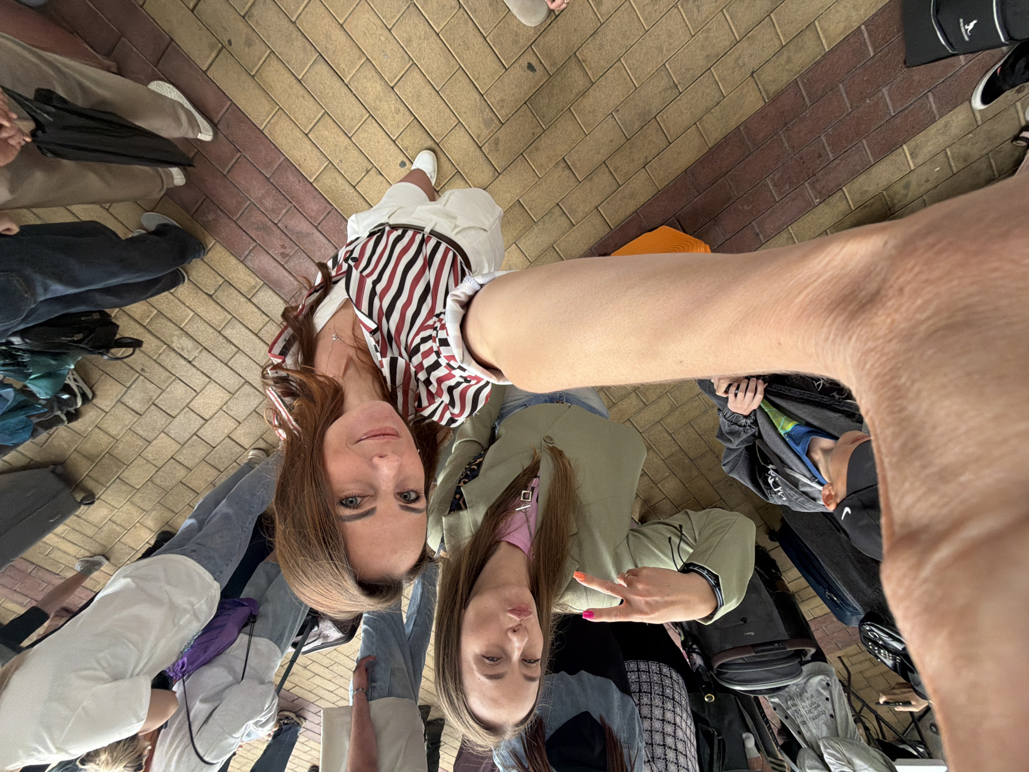 Two women take a selfie outdoors. The woman on the left wears a striped shirt and white pants. The woman on the right wears a green jacket over a pink shirt. The background is a brick walkway. Several other people and bags are also visible in the frame.