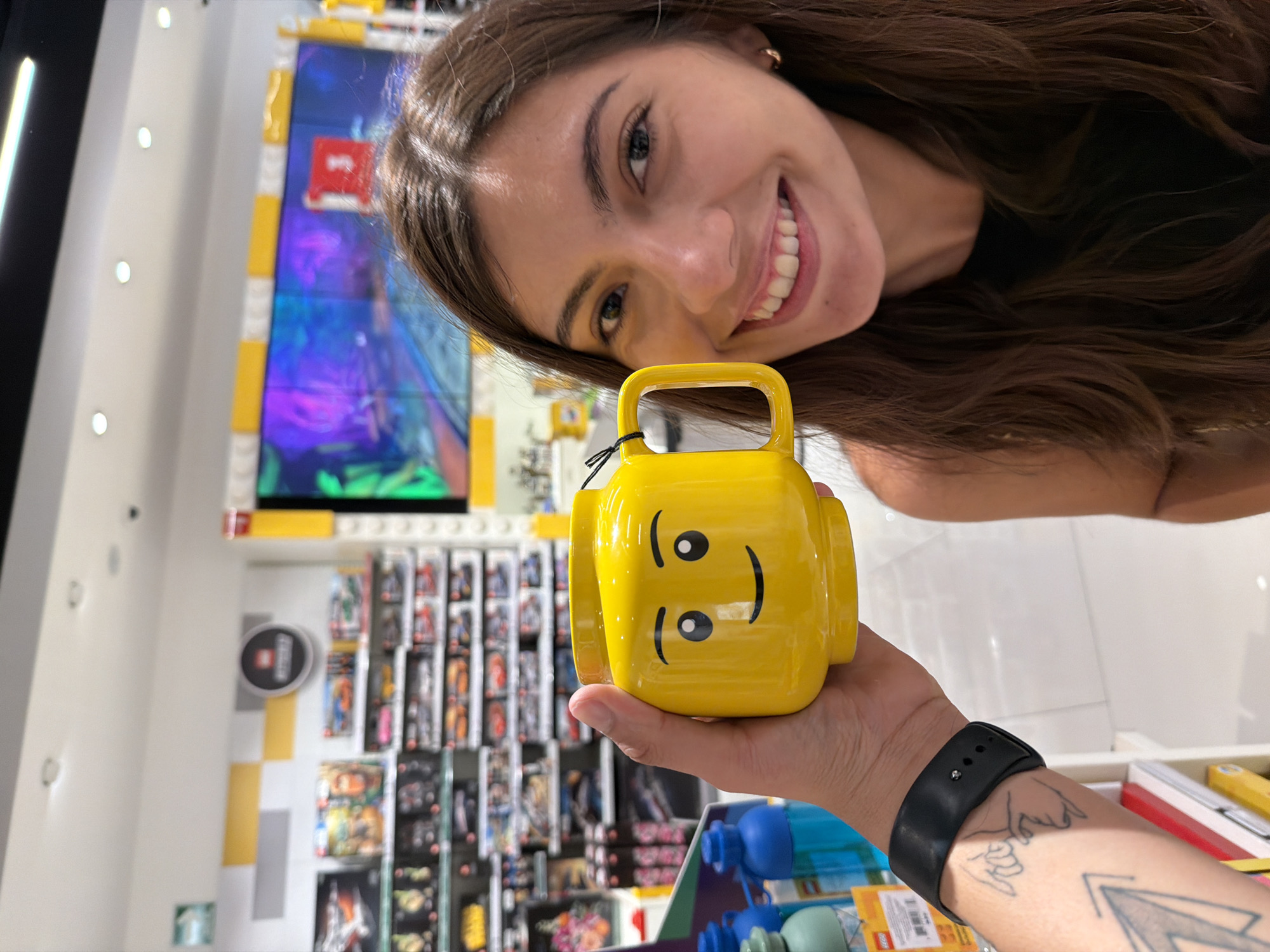A woman with long brown hair smiles at the camera while a hand holds up a yellow, Lego-shaped mug. The scene appears to be inside a Lego store, with shelves displaying Lego sets in the background. Based on the presence of Lego stores in the city, it is possible this photo was taken in Puebla, Mexico.