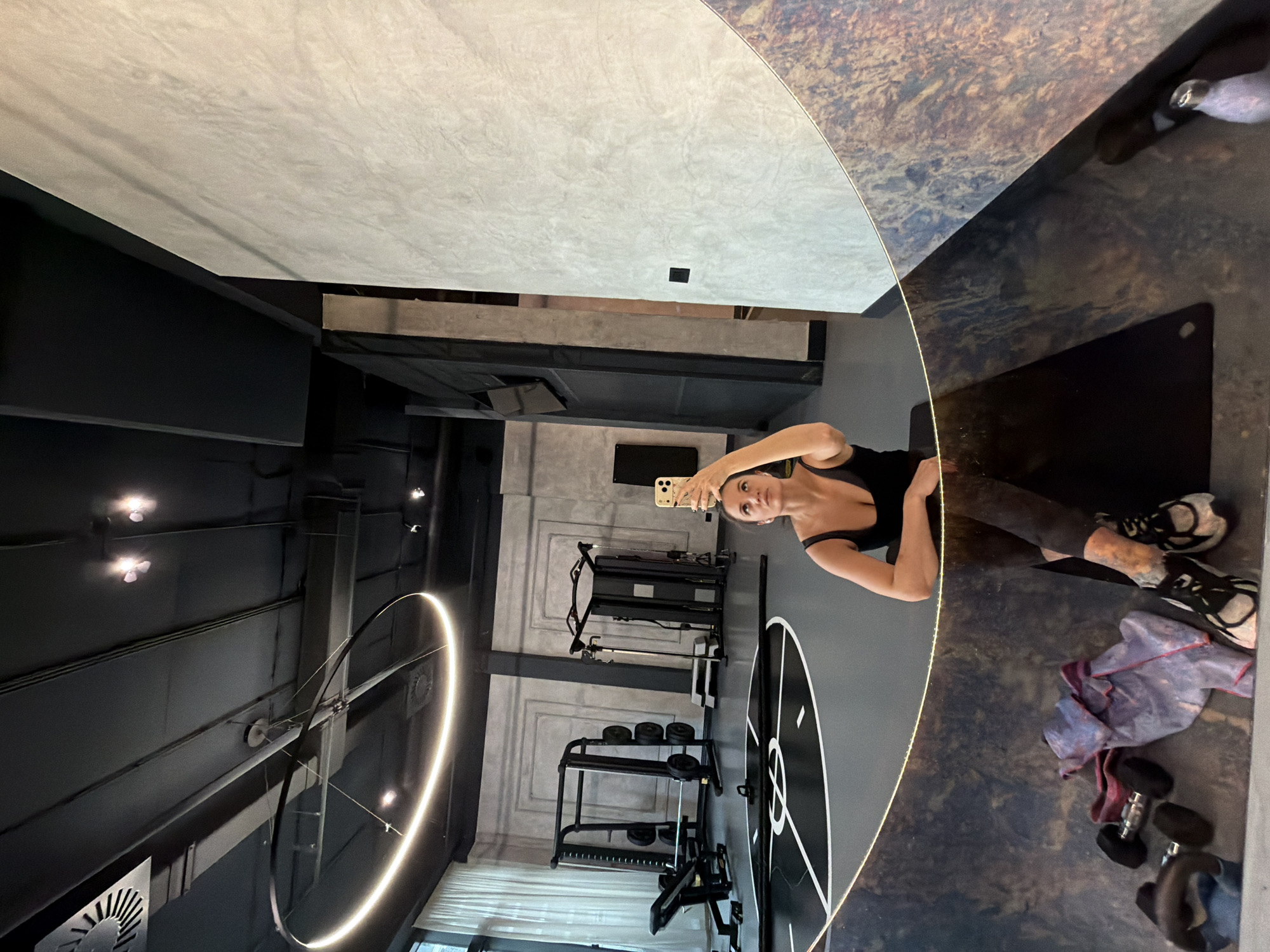 This image captures a mirror selfie taken by an adult female within a modern fitness facility located in Belgrade, Serbia.
**People:**
One adult female is prominently featured, reflected from the chest upwards. She has dark hair, which appears pulled back, and wears a black tank top and dark athletic leggings. Her right arm is raised, holding a light-colored smartphone (possibly an iPhone) above her head to capture her reflection. Her left arm rests on an unseen surface, likely a mat or bench. She is looking upwards towards the phone. Her lower body, including legs and feet clad in athletic shoes (black, white, grey), is visible in the lower portion of the reflection.
**Subjects/Objects:**
The primary subject is the woman taking the selfie. Key objects in the reflected gym environment include:
* **Fitness Equipment:** A black squat rack or power cage with a barbell and several weight plates is visible on the left. Adjacent to it is an adjustable black utility bench. Further right, a multi-station cable machine or similar large black apparatus stands with a grey step platform nearby. A coiled black battle rope rests on the grey floor, extending towards the foreground.
* **Lighting:** A large, prominent circular LED light fixture is suspended from the black ceiling. Additional recessed spotlights are present on the ceiling.
* **Walls:** The walls are textured, primarily light grey, with some sections featuring decorative recessed rectangular panels. A large, plain, light grey concrete-textured wall is visible on the right. A large window with white sheer curtains is on the far left.
* **Floor:** A dark grey floor features distinctive white circular and linear markings, suggesting designated zones for exercises or agility drills.
* **Foreground (reflected):** In the bottom left of the reflection, two black dumbbells, a folded purple and blue towel, and a black exercise mat are visible on the floor. A glimpse of a clear object, possibly a water bottle, is on the far right.
* **Ceiling:** The ceiling is black, with exposed ventilation ducts and other architectural elements.
**Actions/Interactions:**
The woman is actively engaging in taking a selfie photograph using her smartphone, positioning it to capture her reflection in the large mirror.
**Environment:**
The scene is an indoor, contemporary fitness studio or gym with an industrial-minimalist design aesthetic. The color palette is dominated by black, grey, and white, with contrasting illuminated elements. The facility appears well-maintained and organized. While the city is identified as Belgrade, Serbia, a specific street address or the name of the fitness establishment cannot be determined from the visual information provided.