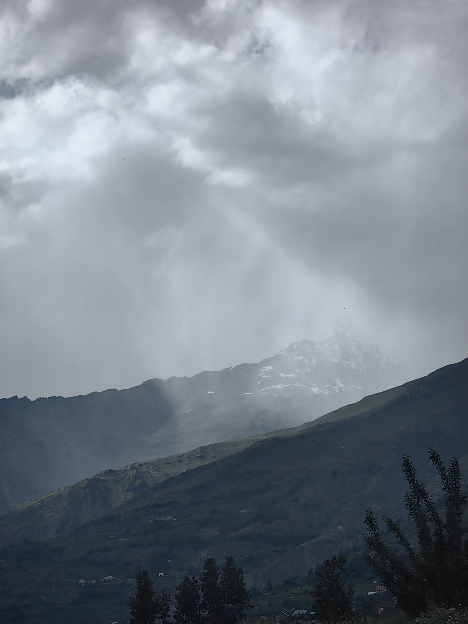 The image shows a mountainous landscape with a cloudy sky. In the foreground, there are dark green hills with some houses and trees visible at the bottom. The middle ground features rolling hills covered in vegetation, with some patches of lighter green. In the background, a tall, snow-capped mountain peak emerges from the clouds, with rays of light breaking through the overcast sky. The overall atmosphere is dramatic and moody, with a sense of vastness and natural beauty. The scene is likely located in the city of Tandi, India, given the mountainous terrain and vegetation characteristic of the region.