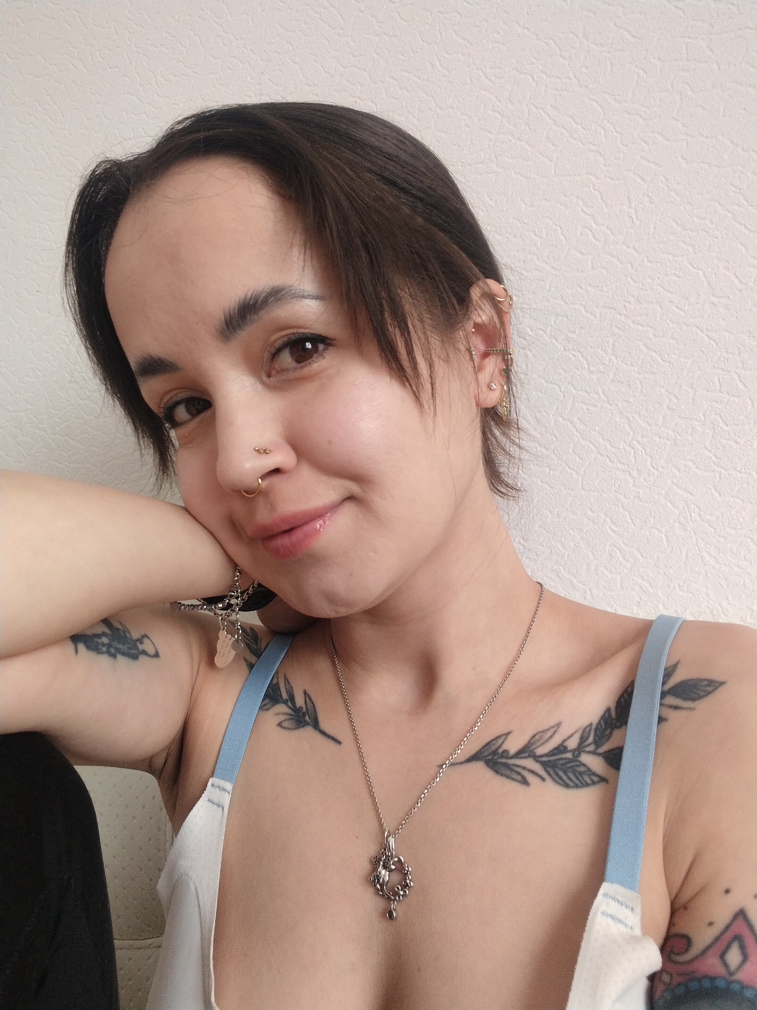 A selfie of a fair-skinned person with short, dark hair, resting their cheek on their hand. The person is wearing a white tank top with light blue straps. Visible tattoos include a leafy branch across the chest, another on the upper arm near the shoulder, and a partial one on the lower arm. The person is wearing a necklace, earrings, and nose rings. The background is a plain, light-colored wall. No location in Noviy, Russia is evident.