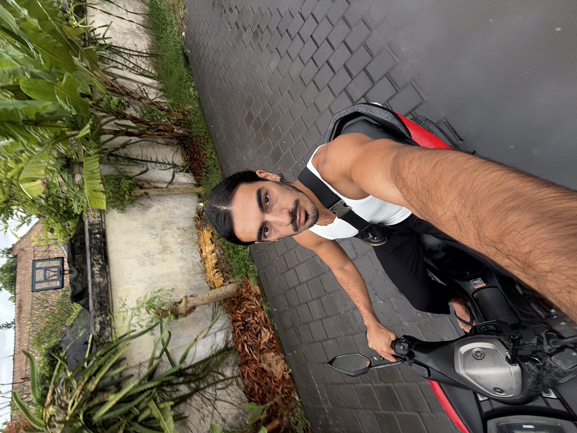 The photograph, taken in what appears to be North Kuta, Indonesia, shows a man on a red and black motorcycle. He is fair-skinned with dark hair pulled back, a mustache, and a goatee. He wears a white tank top, dark pants, and flip-flops. A black strap crosses his chest diagonally. The background includes a brick road, tropical vegetation including banana trees and aloe plants, and a wall with a basketball hoop. The road appears wet, suggesting recent rain. The man is looking directly at the camera, holding the motorcycle handlebars.