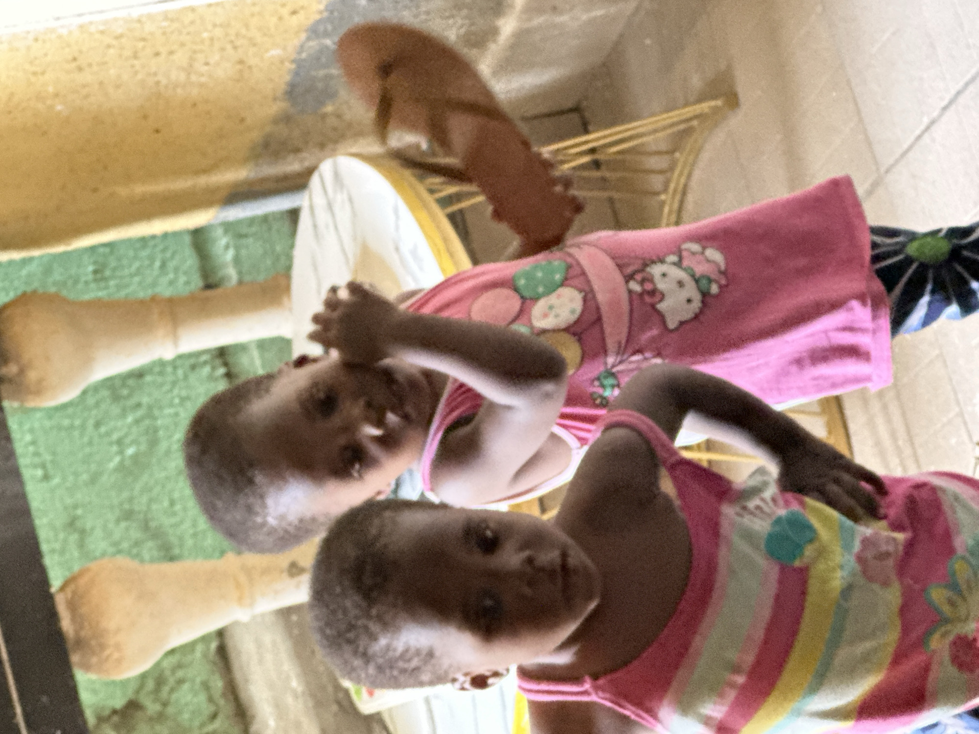 In Narayi, Nigeria, two young girls stand in an indoor setting. The girl on the left, with short dark hair, wears a pink tank top adorned with horizontal stripes of pink, yellow, and white, and a floral pattern. The girl on the right, also with short dark hair, is dressed in a pink dress featuring a Hello Kitty graphic and a pattern of colorful balloons. Her right hand is raised, and she holds a brown flip-flop. The background shows elements of an indoor environment, with textured walls, pillars, and furniture.
