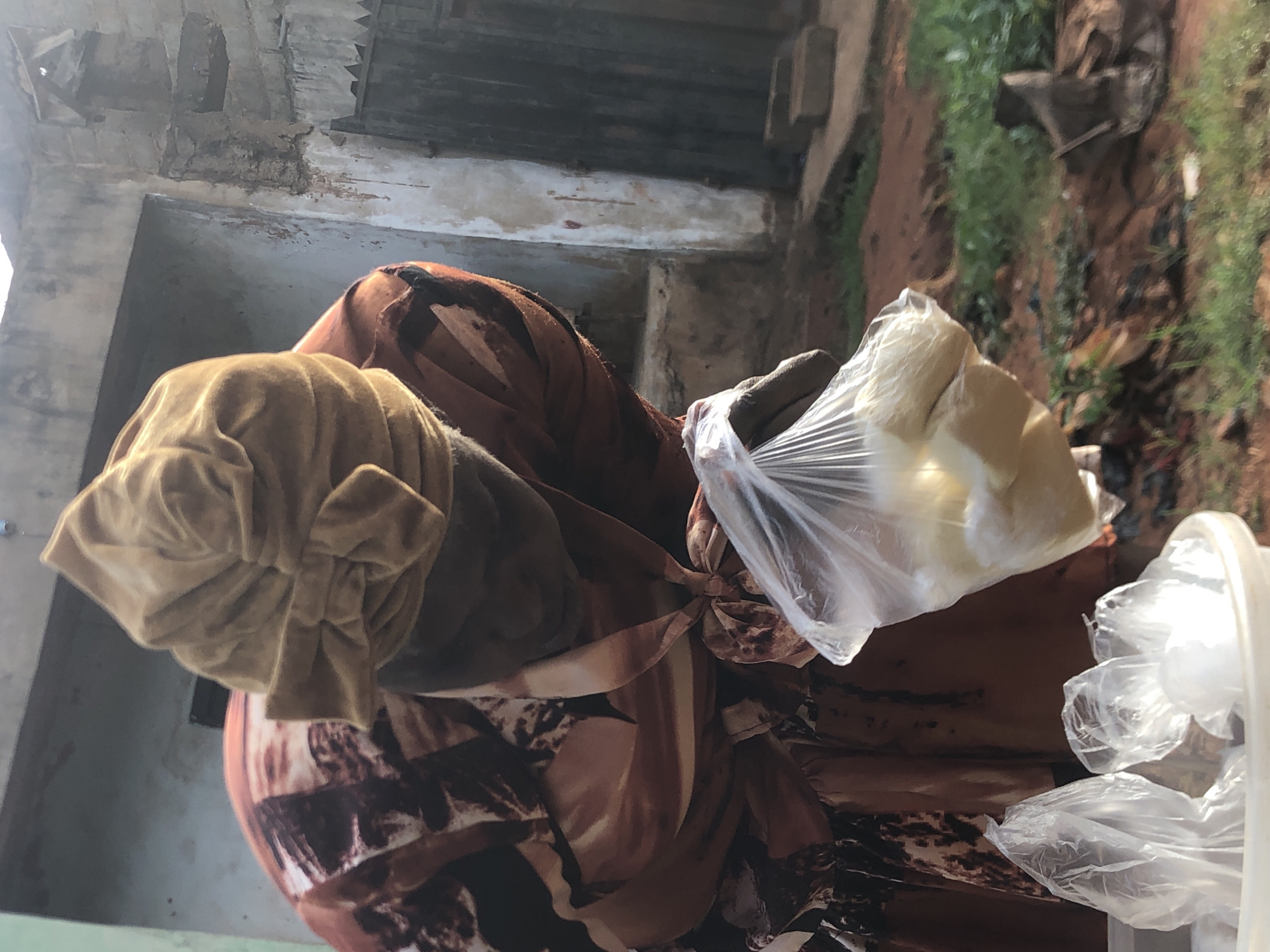 A person in Ukwu-Abwa, Nigeria, wearing a tan-colored head covering and a brown patterned garment, holds a clear plastic bag filled with white, solid objects. The person is standing in front of a building with a weathered, deteriorating facade. The environment includes green vegetation and reddish-brown soil in the background.