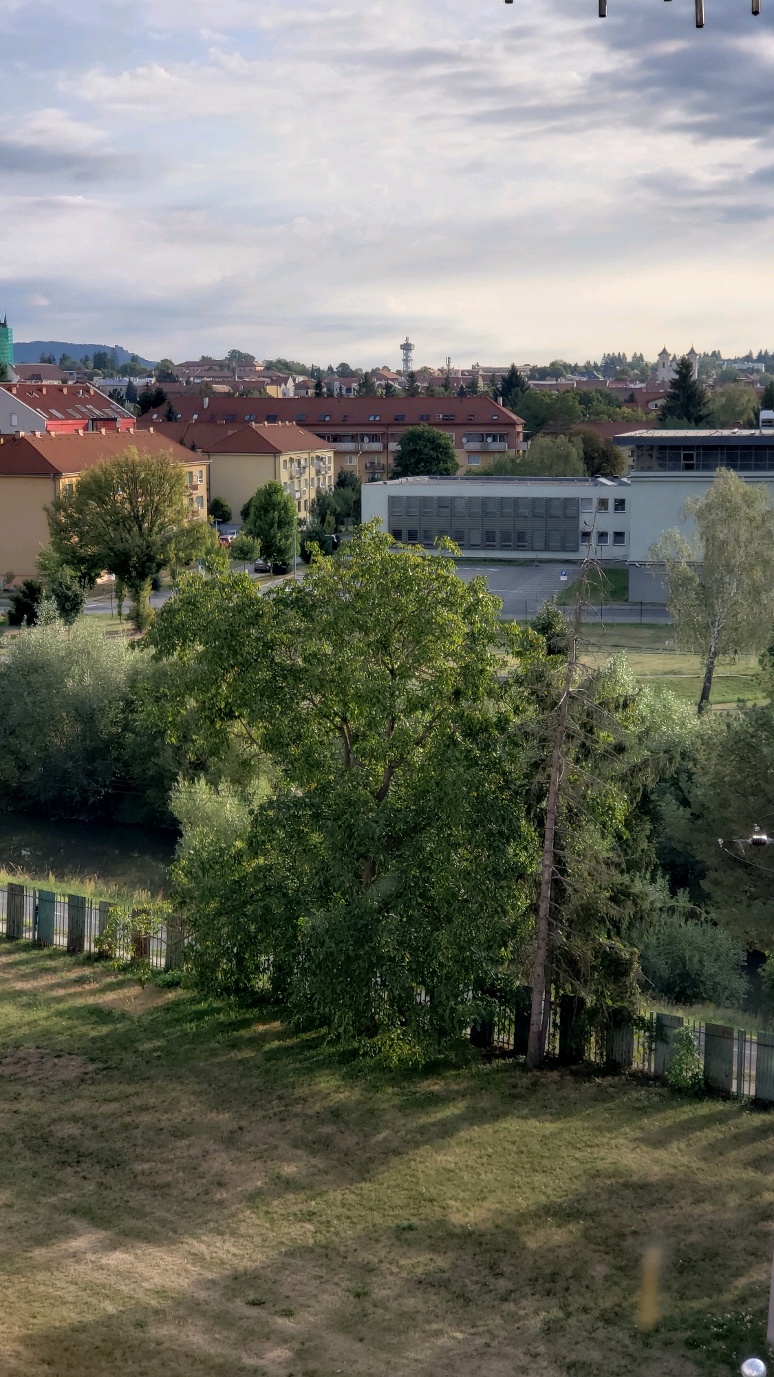 The image displays a panoramic view of a cityscape, likely Prešov, Slovakia, under a partly cloudy sky. In the foreground, a grassy lawn slopes gently downwards, casting long shadows from unseen trees. A dark metal fence runs along the bottom edge of the lawn, separating it from a body of water.
Beyond the lawn, lush green trees dominate the midground, with a large deciduous tree in the center creating a significant focal point. To the left of this tree, a small pond or narrow river is visible, reflecting the greenery. Behind the trees, the middle distance reveals residential buildings with red-tiled roofs, characteristic of European architecture. Further back, a modern, light-colored building with a grid-like facade stands out against the skyline.
In the background, the city of Prešov unfolds with a mix of smaller houses and taller structures. A prominent tower with an antenna structure is visible on the horizon, and a church steeple can be spotted to the right. The overall impression is one of a residential area adjacent to a green space, with the city stretching out beyond. No people are visible in the image.