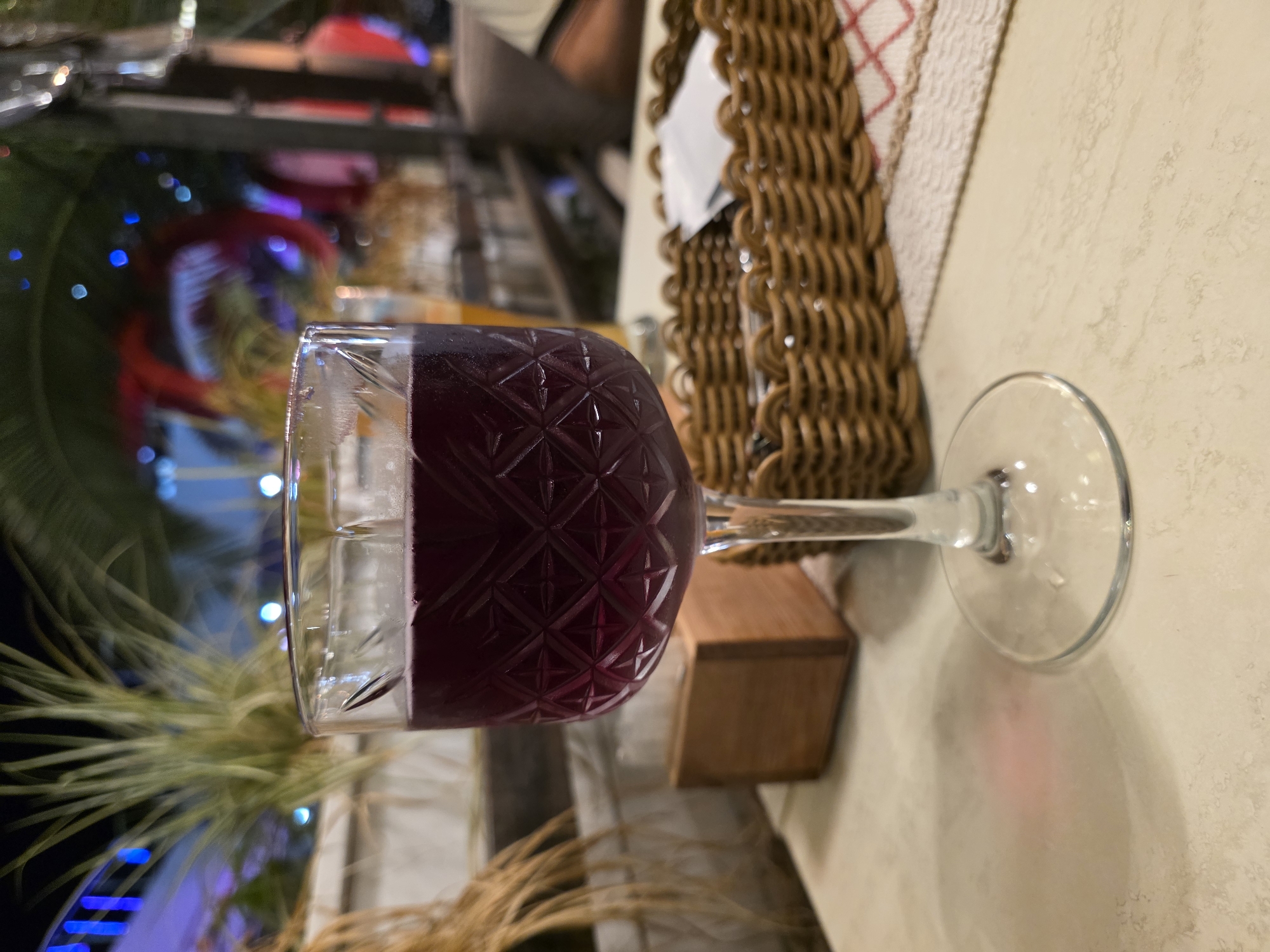 A close-up, eye-level shot shows a stemmed glass filled with a dark purple beverage, likely wine, on a light-colored, textured table. The glass has a diamond-patterned etching on its bowl. In the background, out of focus, are palm fronds and blurred colorful lights, suggesting an outdoor evening setting. To the right of the glass is a woven basket containing what appears to be sugar packets and napkins. A small wooden box is visible behind the glass to the left. The overall impression is of a relaxed evening at an establishment in Sochi, Russia, possibly a restaurant or bar.