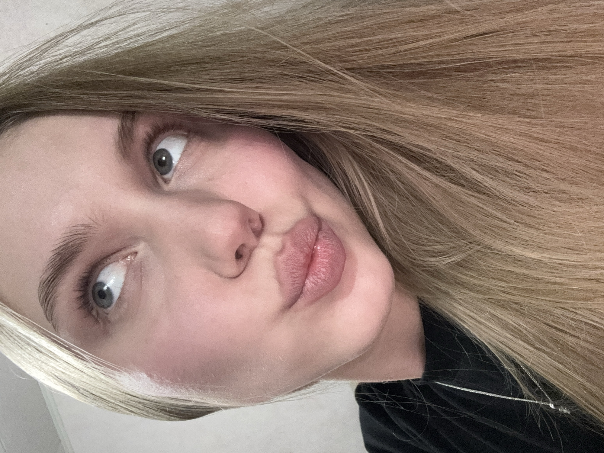 A close-up portrait depicts a young individual, likely female, in an indoor setting identified as the city of Perm, Russia. The subject has fair skin, long light blonde hair flowing downwards and to the right, and light blue-grey eyes looking upwards and to the right. Her lips are distinctly pursed in a pouting or kissing gesture. Visible facial features include well-defined, light brown eyebrows, long eyelashes, and subtle redness on the cheeks. She wears a black garment and a thin silver-colored chain necklace with a small, indistinct pendant. The background is an out-of-focus, uniformly light-colored wall.