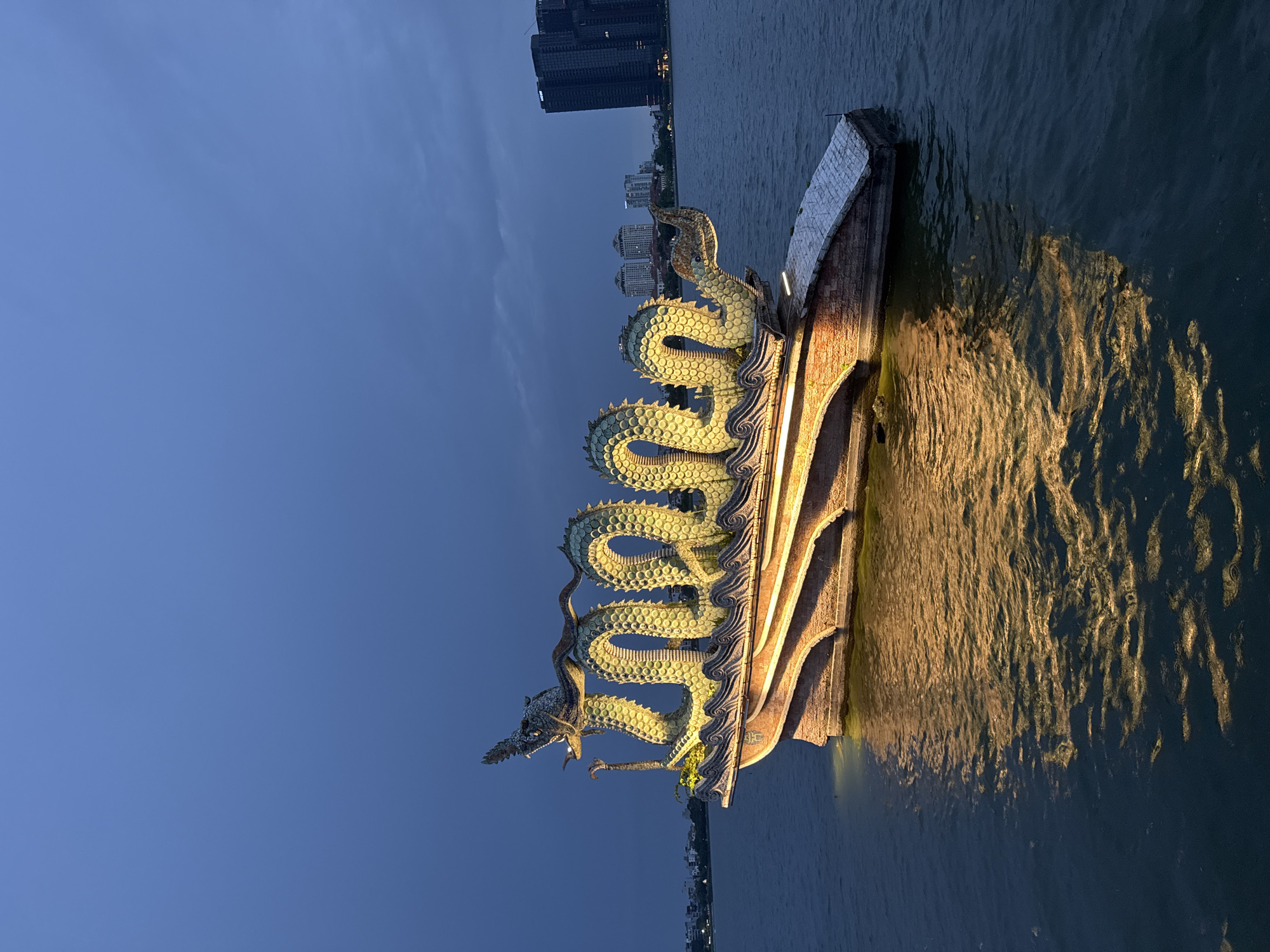 The image captures a nighttime view of a dragon boat floating on a body of water, likely the West Lake in Hanoi, Vietnam. The dragon boat is elaborately decorated with a green, segmented dragon sculpture that wraps around its length. The dragon has a detailed head with a crest and sharp teeth. The boat itself is constructed with a reddish-brown brick base and features tiered, curved sides. Warm yellow lights illuminate the underside of the boat and the dragon sculpture, creating a vibrant glow that reflects on the dark water. In the background, the skyline of Hanoi is visible, featuring several modern high-rise buildings and some smaller structures, all silhouetted against a dusky, twilight sky with subtle cloud formations. The water is calm, with gentle ripples distorting the reflections of the lights. The overall scene is one of illuminated artistry against a darkening urban backdrop.