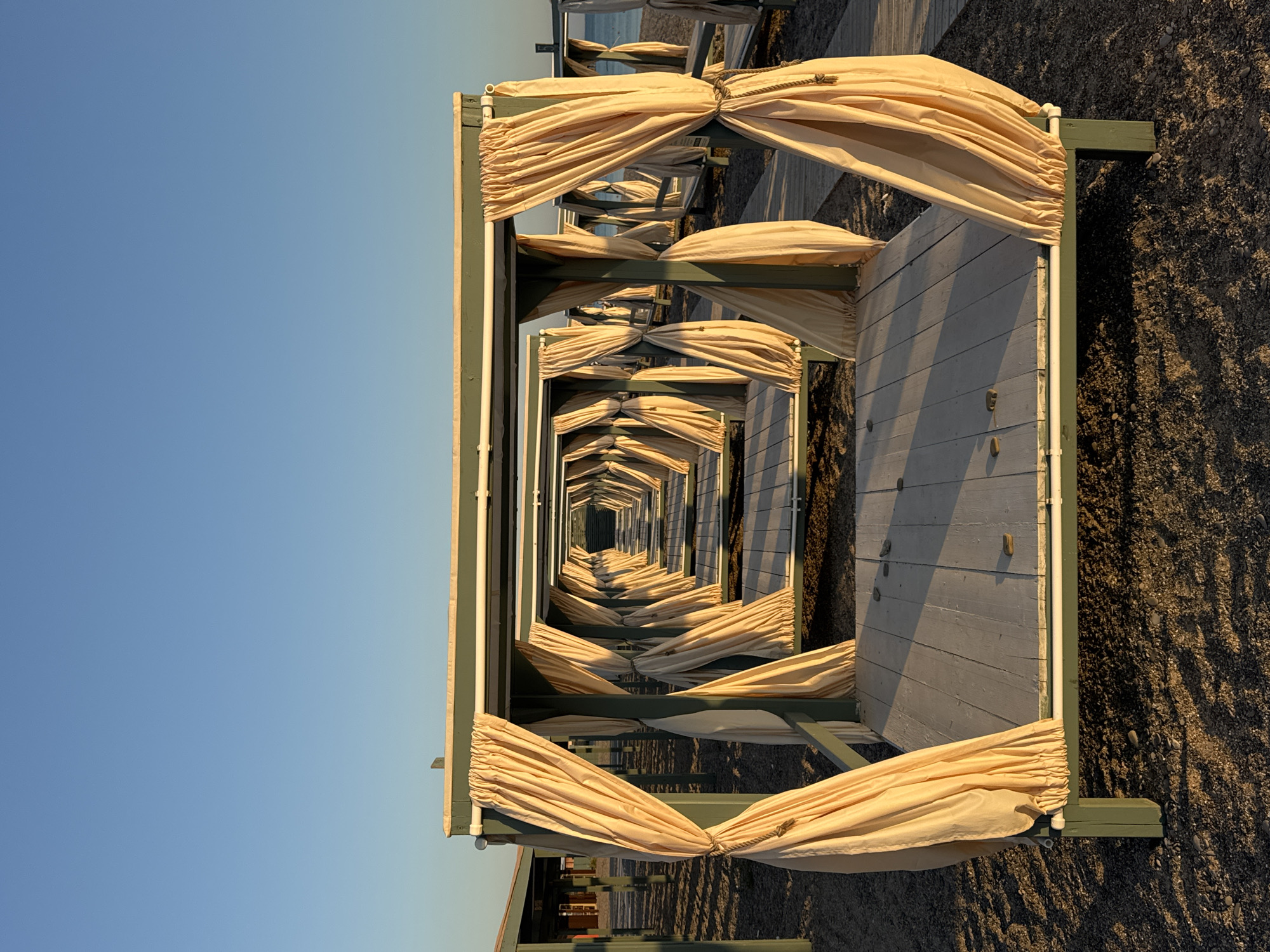 The image depicts a line of open-air cabanas on a beach in Novofedorovka, Ukraine. The cabanas are constructed with green wooden frames and feature light yellow curtains that are drawn back with ropes, revealing wooden decks inside. The perspective creates a strong sense of depth, with the cabanas receding into the distance and appearing to form a tunnel-like effect. The ground is covered in dark pebbles, and a wooden walkway is visible to the right of the central cabanas. The sky is clear and blue, suggesting a sunny day, and the lighting indicates it is either early morning or late afternoon. There are no people visible in the image, and the cabanas appear unoccupied. The scene suggests a resort or recreational area on the coast.