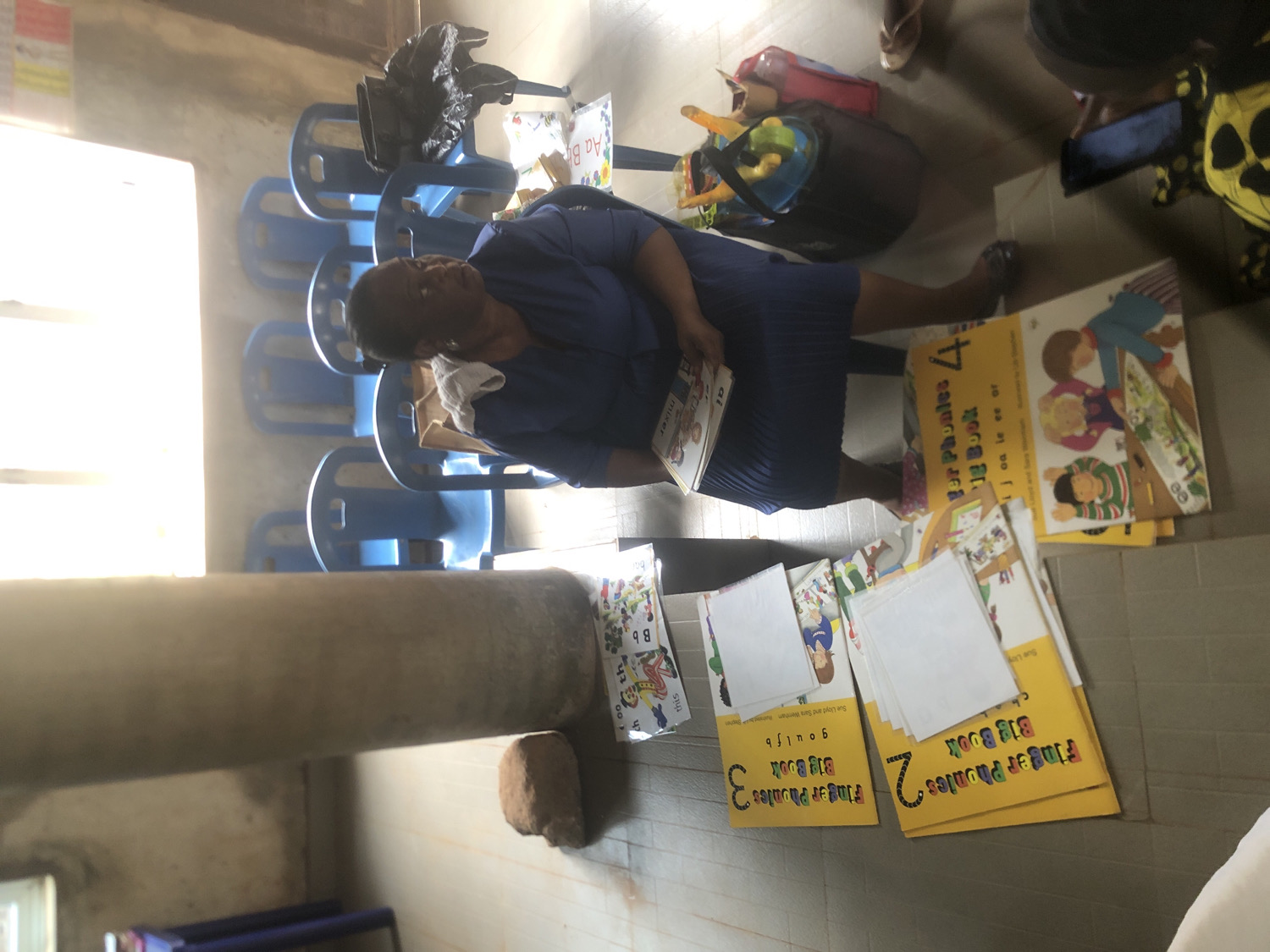 The image depicts an indoor scene, likely a classroom or educational space. An adult woman, dressed in blue, is seated or crouching on a light-colored tiled floor, holding an open book. Around her, several educational books, predominantly from the "Finger Phonics Big Book" series, are spread out on the floor. These books are numbered 2, 3, and 4, indicating different levels, and also feature children's illustrations.
The activity appears to be an educational session, possibly a reading or phonics lesson, or preparation for one. The woman seems focused on the reading material. In the background, several blue plastic chairs are arranged. A large, grey cylindrical pillar stands in the mid-ground, and bright light streams in from a window on the left side of the frame. Other objects include a black bag and a red/black bag containing toys, suggesting the presence of children or young learners. A small, reddish brick or rock is also visible on the floor near the pillar.
Visible text on the book covers includes "Finger Phonics Big Book", along with numbers "2", "3", and "4". Author names "Sue Lloyd and Sara Wenham" are also legible. Specific phonics sounds and words such as "ch", "goulfb", "ijoa le ee or", "mixer", "this", "Aa Bb", "th", and "oo" are printed on the books.