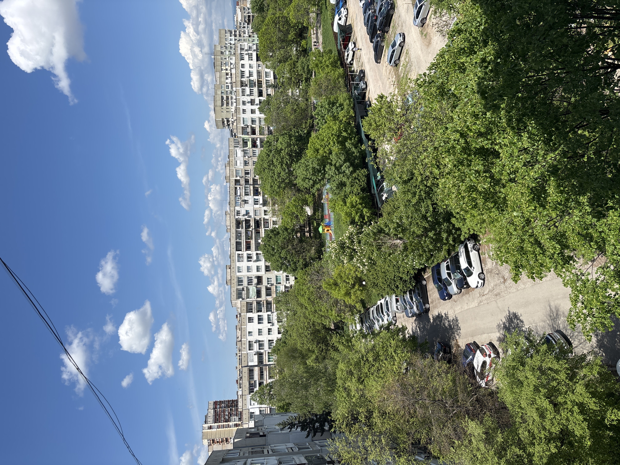 The image presents an elevated view of an urban landscape, likely in Sofia, Bulgaria, characterized by apartment buildings and ample greenery. The left side of the frame features a blue sky dotted with cumulus clouds, contrasting with the densely populated area on the right. Several multi-story apartment buildings with repeating window patterns are visible, typical of Soviet-era construction. The area is interspersed with mature trees, some obscuring the buildings and others lining streets and parking areas. Cars are parked along the road and in designated parking spots. There is a playground among the trees, featuring colorful play equipment. Electrical wires are visible against the sky in the lower left corner. The scene appears to be captured during daylight hours, with shadows cast by the buildings and trees. There are no people depicted in this image.