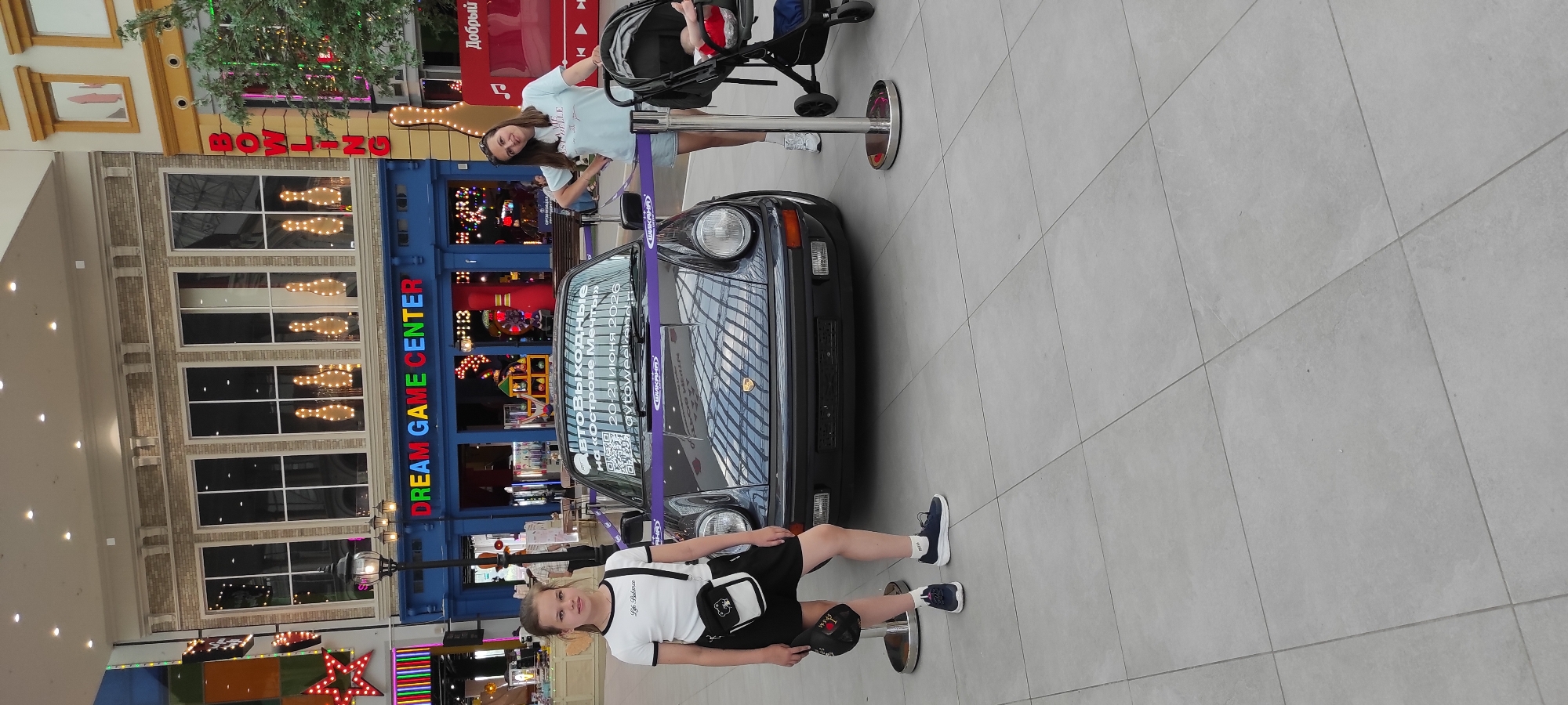 In Moscow, Russia, two girls and a baby are in front of a dark gray Porsche parked indoors. The Porsche is on display, cordoned off by a purple ribbon. The backdrop features a building with large windows displaying bowling pins lit with bulbs. A sign above the windows reads "DREAM GAME CENTER" in multi-colored letters. The floor is tiled with light gray square tiles. The girl on the left is wearing a white t-shirt, black shorts, and white socks with blue sneakers, and is holding a black hat. The girl on the right is wearing a light blue t-shirt and denim shorts, with white sneakers. A baby in a stroller is positioned to the right of the Porsche.