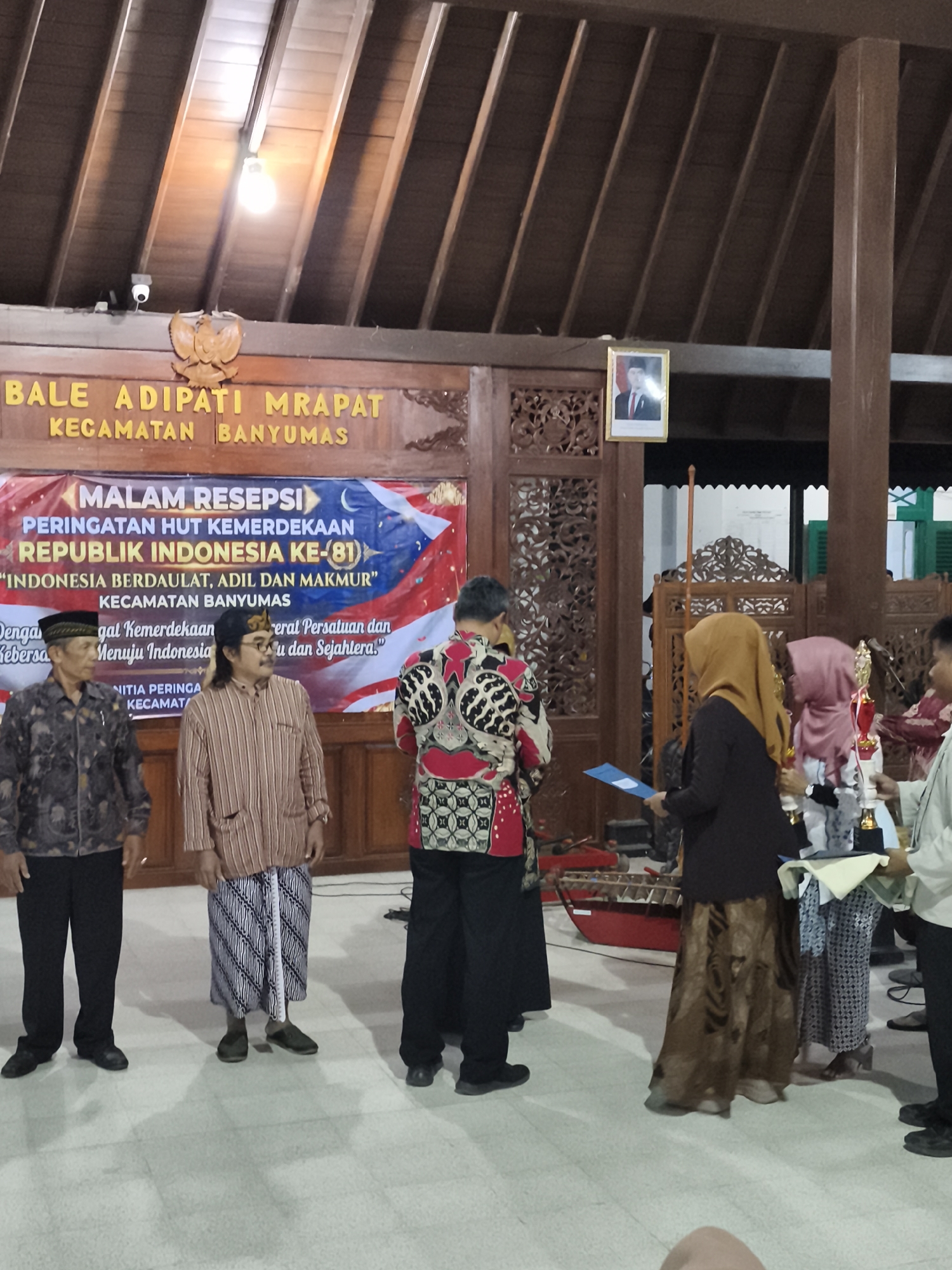 In a wooden hall in Banyumas, Indonesia, a group of people are gathered for what appears to be an awards ceremony. The hall features a high, sloped wooden ceiling with exposed beams and decorative carvings. A banner hangs across the back wall, featuring Indonesian text that translates to "Republic of Indonesia Independence Day Celebration". The banner also displays the slogan "Indonesia Sovereign, Just and Prosperous".
In the foreground, several men are dressed in traditional Indonesian attire, including batik shirts and sarongs. One man on the far left wears a dark patterned shirt and black pants, while the man next to him sports a striped shirt and a batik sarong. A third man, partially visible on the right, wears a red and black patterned shirt with black pants.
To the right of the men, several women are also present. One woman in a brown hijab and a long patterned skirt holds a blue certificate or folder. Another woman, wearing a pink hijab, stands beside her, holding a trophy. A third woman, mostly obscured, appears to be receiving an award.
In the background, a portrait of a man in a suit and tie is displayed on the wall. A wooden musical instrument, possibly a gamelan, is visible on the floor. The overall atmosphere suggests a formal cultural event celebrating Indonesian independence.