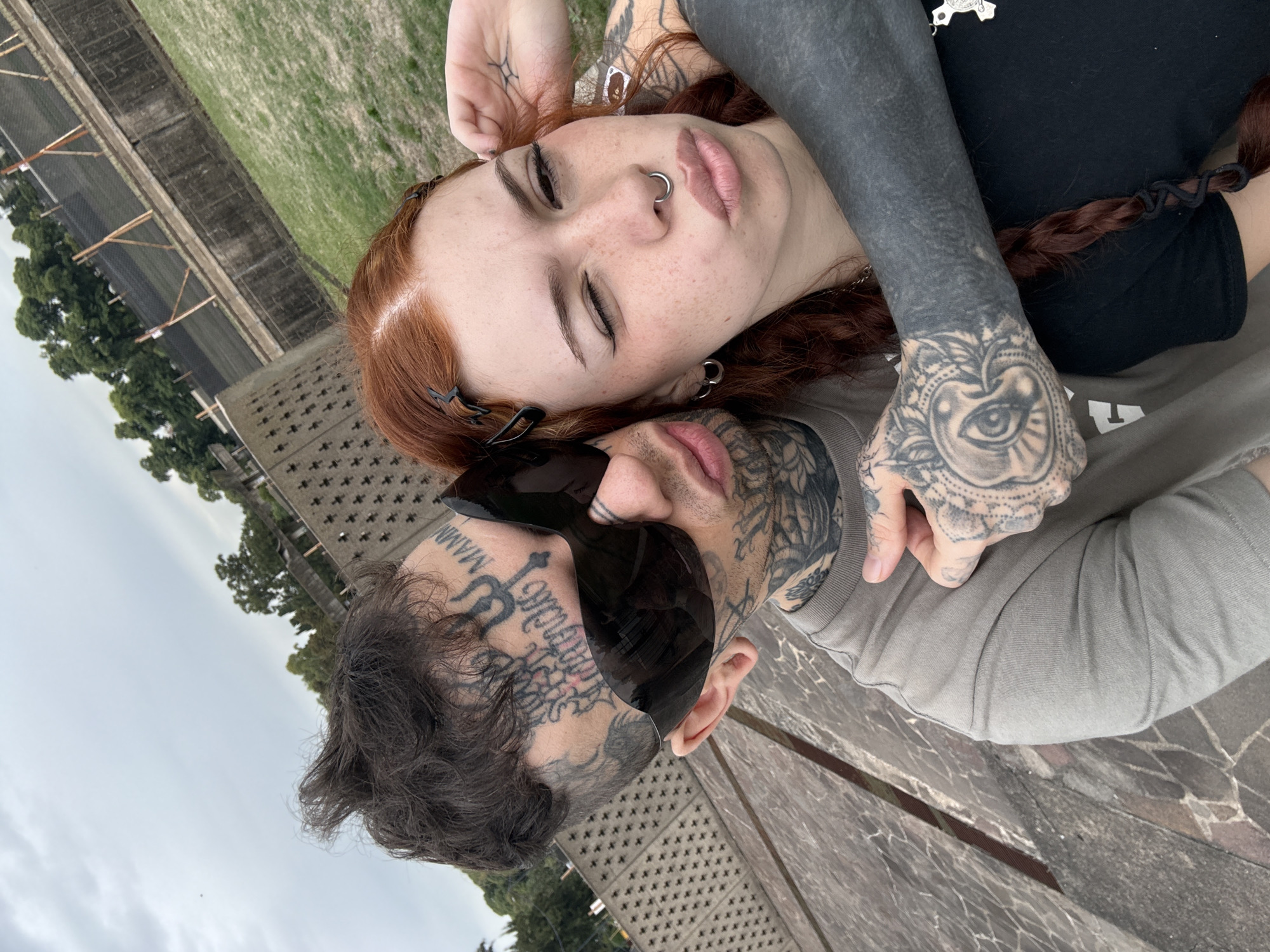 A close-up, angled outdoor portrait features two heavily tattooed individuals, an adult male and an adult female, in a close embrace. The scene is located in the Recoleta neighborhood of Buenos Aires, Argentina, near structures consistent with the Facultad de Derecho or the Plaza de las Naciones Unidas, indicated by a distinctive textured concrete wall with a grid pattern in the background.
The male, positioned above and embracing the female, has short, dark, disheveled hair. His face is heavily tattooed with script and symbols, including "MAMMY" visible on his right temple and a trident-like design near his left eye. He wears large, dark, wrap-around sunglasses that obscure his eyes and upper nose. His visible left arm and hands are also extensively tattooed. He is dressed in a muted olive green or grey short-sleeved t-shirt.
The female is positioned below the male, leaning into him with her eyes closed, displaying a relaxed expression. She has long, red/auburn hair, partially held back by dark hair clips on her right side, with a visible braid on her left shoulder. Her face is covered in numerous freckles. She has a silver septum piercing and silver hoop earrings. Her arms are heavily tattooed, with her right forearm featuring a blackout tattoo and her visible left hand displaying intricate blackwork, including an eye motif. She wears a black short-sleeved t-shirt. Her left hand is raised near her temple.
The background includes a light-colored, brutalist-style concrete wall with a repeating pattern of recessed square or cross shapes. Beyond this wall, green foliage from trees is visible against a light grey, overcast sky. The ground to the lower left consists of an irregularly paved surface, and a grassy area is visible in the mid-ground.