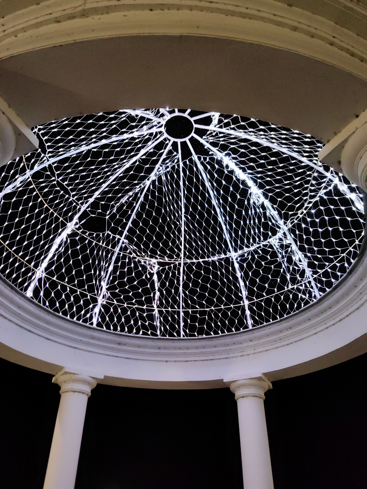 An upward view captures a white domed structure at night. The dome's opening reveals a black sky filled with a lattice of white lights forming a geodesic pattern, resembling a net or a dome of stars. The outer structure of the dome is a white, circular architectural element, supported by two white pillars visible in the foreground. The image appears to be taken from a ground-level perspective, looking up through the opening of the dome. The scene is likely a decorative feature, possibly a gazebo or part of a building, illuminated for nighttime viewing. This scene is located in Myadzyel, Belarus.